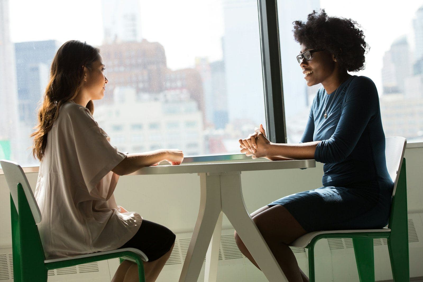Two women talking across a small table by a bright window
