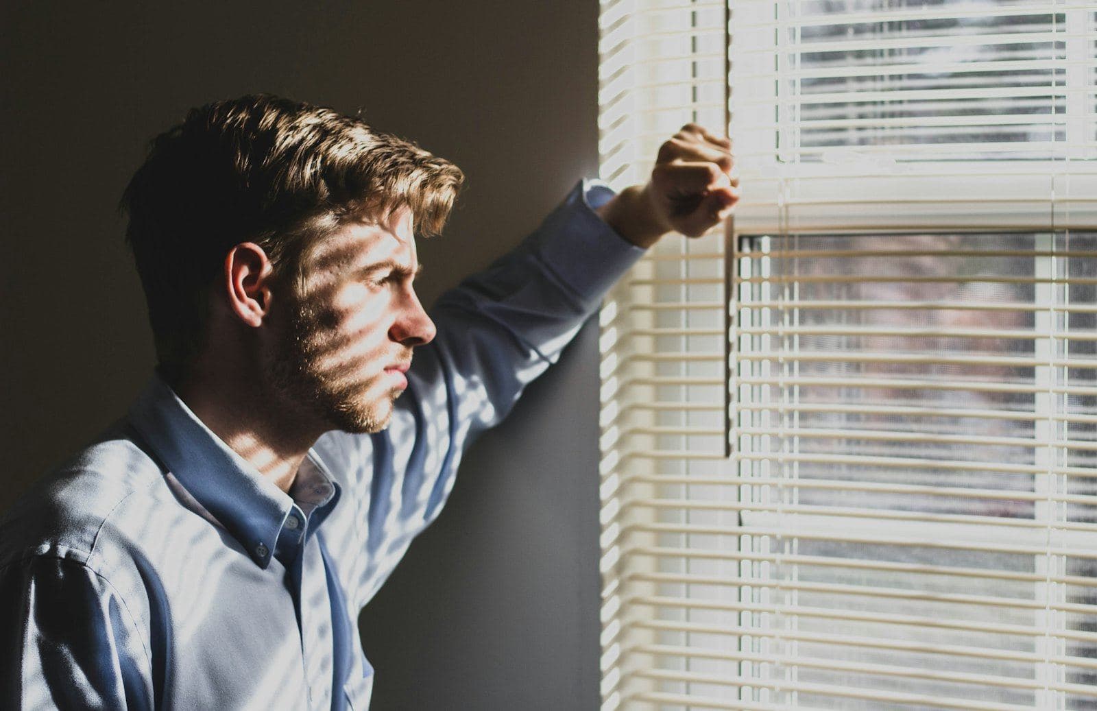 A man sitting by a window with the blinds half open, deep in thought