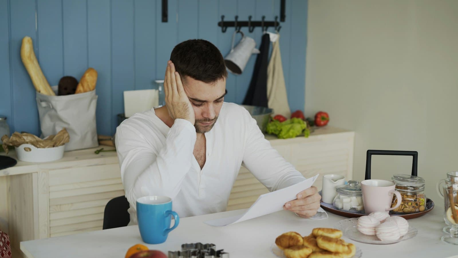 A man at a kitchen table reading a letter, his hand pressed to the side of his head