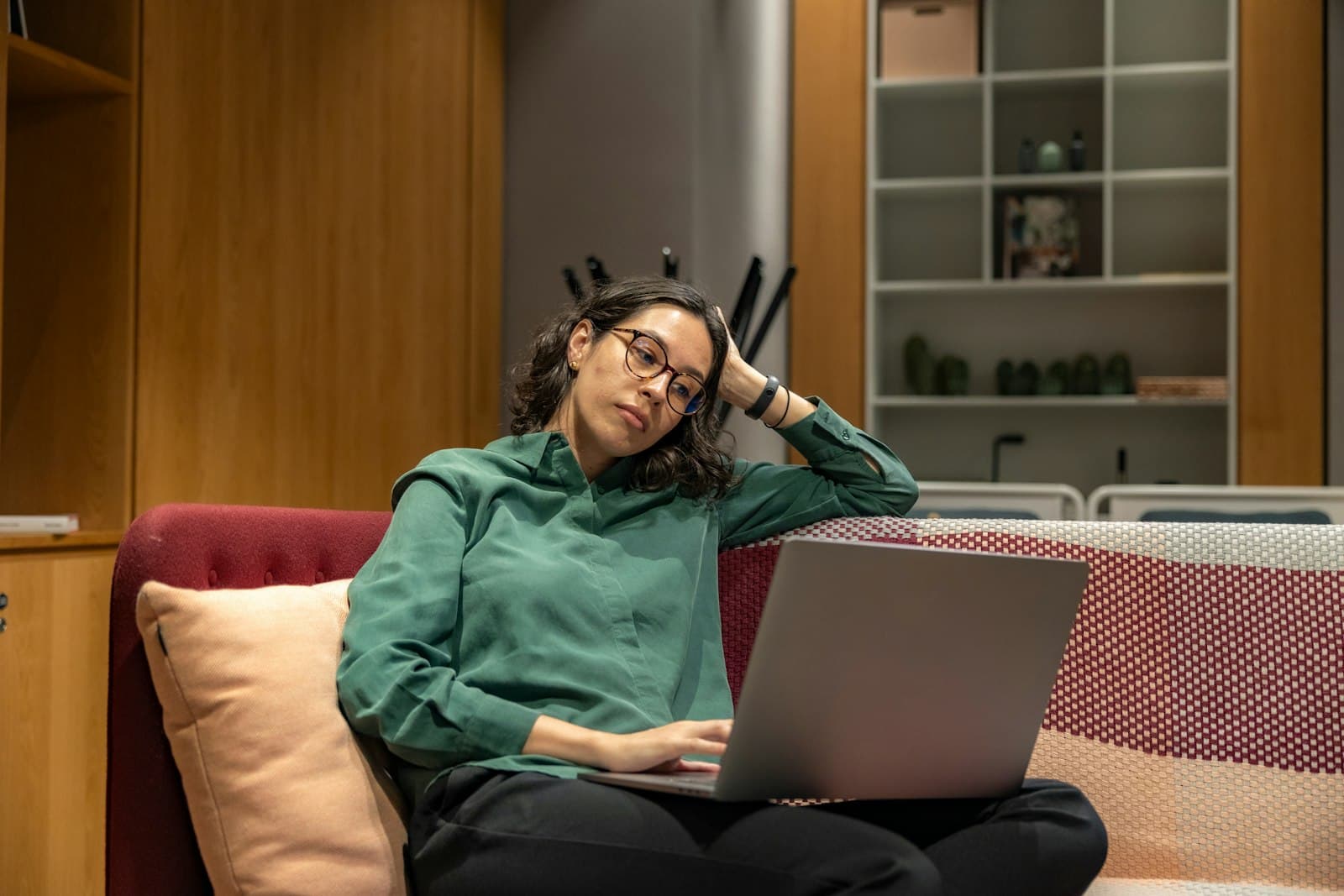A woman in glasses on a sofa with a laptop, resting her head on her hand with a frustrated look