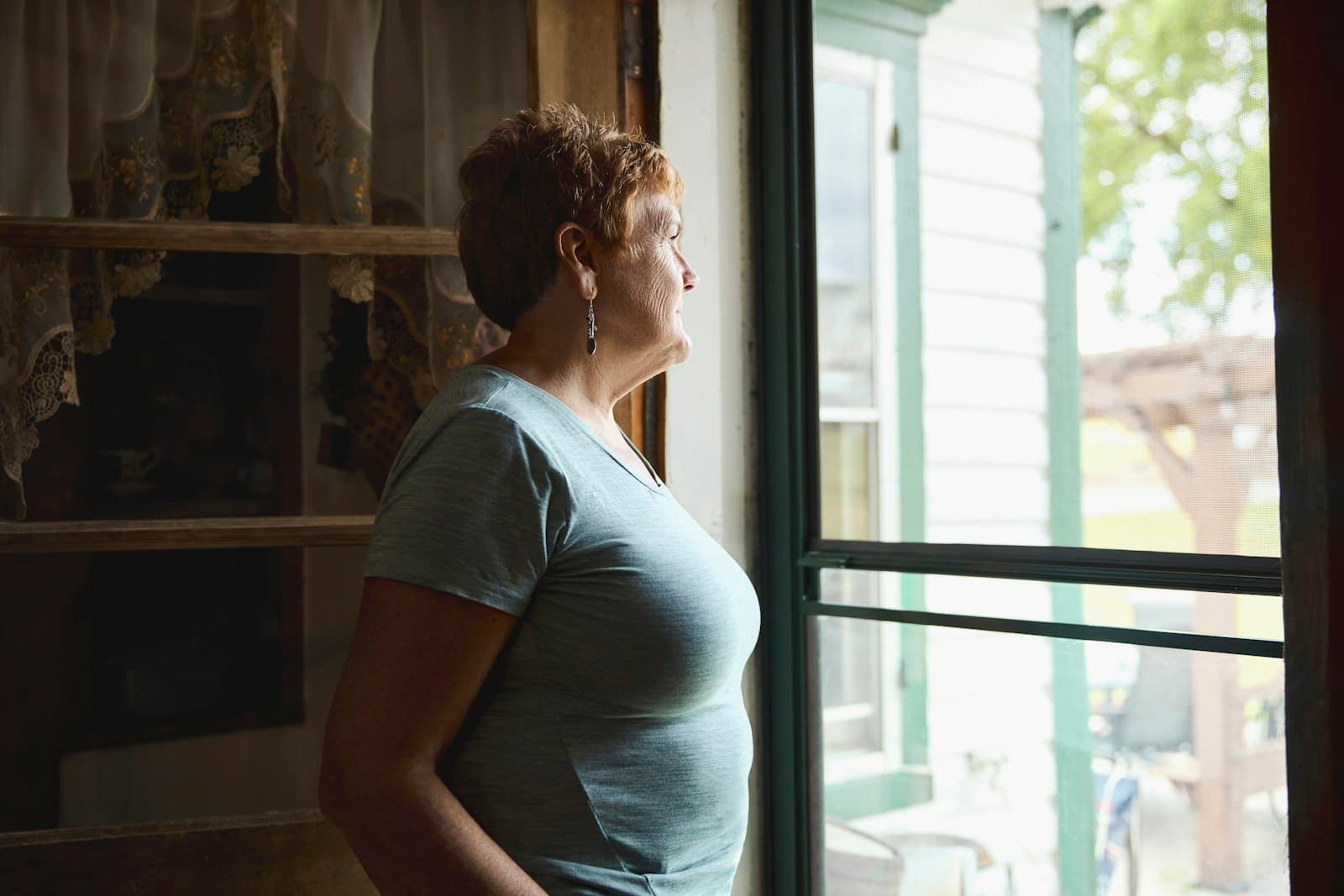 An older woman standing at a screen door, looking out at the yard