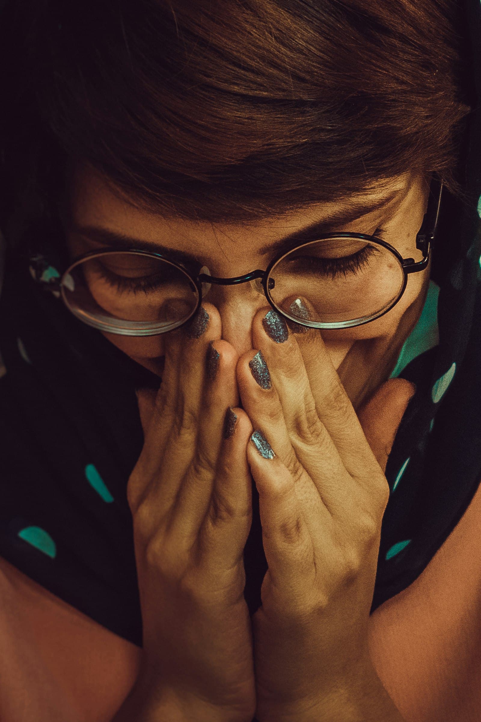 A woman in round glasses pressing her hands to her face, eyes lowered