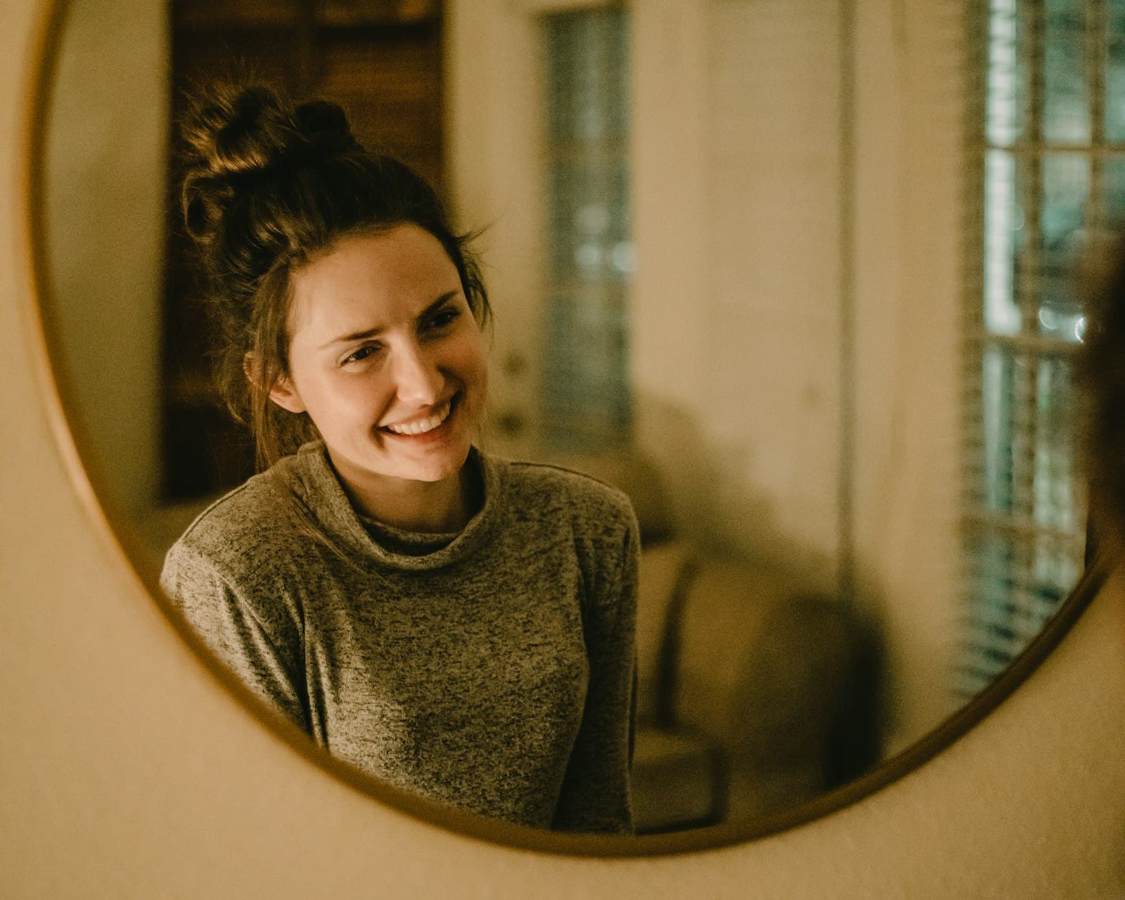 A woman in a grey turtleneck smiling at her reflection in a round mirror