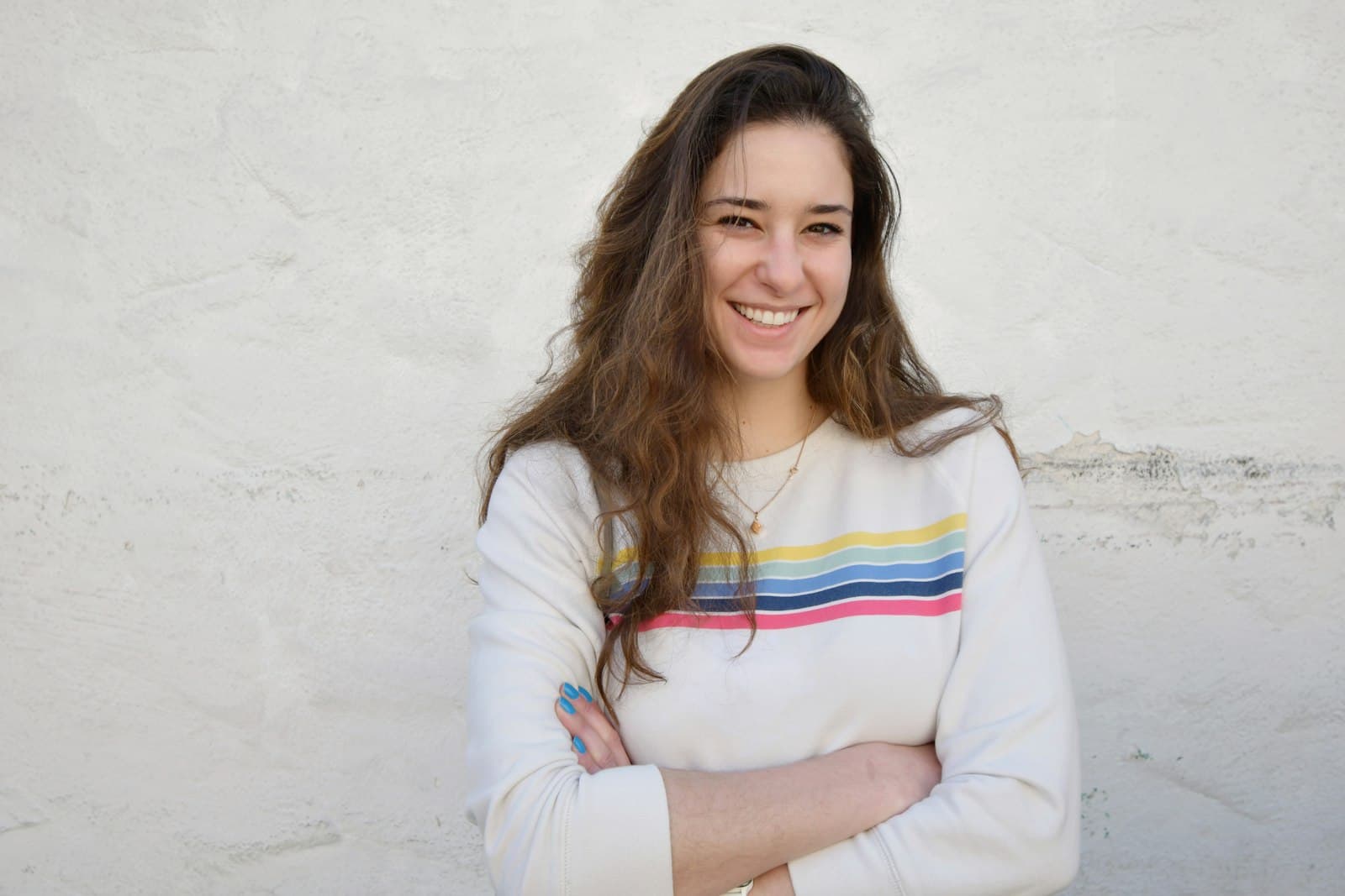 A smiling woman in a striped sweater standing with her arms crossed against a white wall