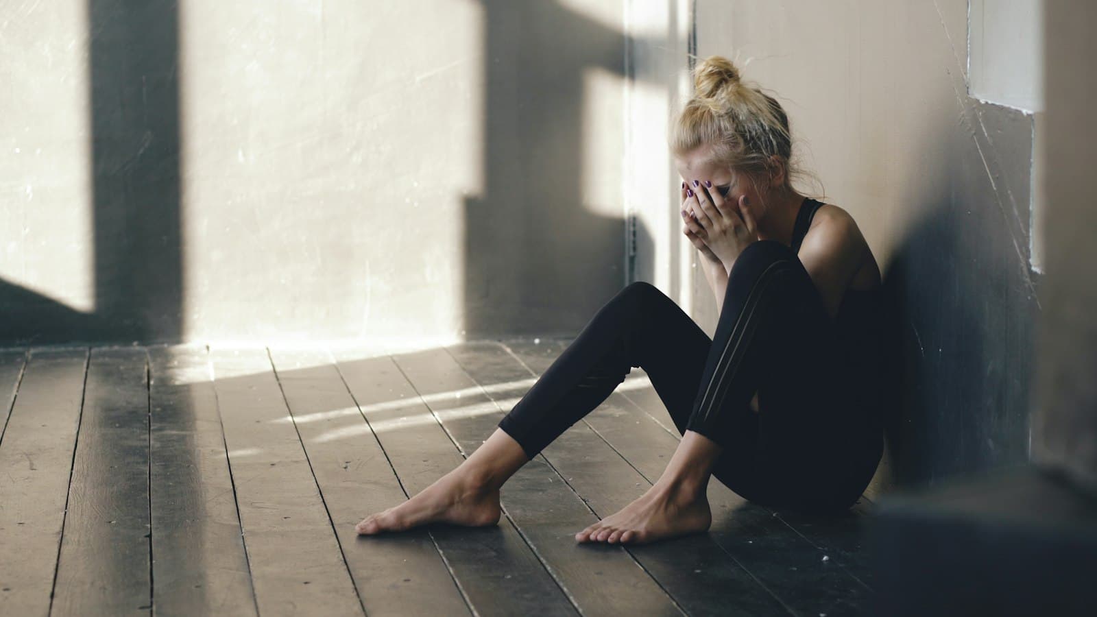 A woman sitting on a wooden floor against a wall, covering her face with her hands