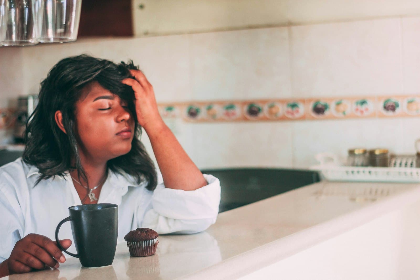 A tired woman at a kitchen counter with a mug, her hand in her hair and eyes closed