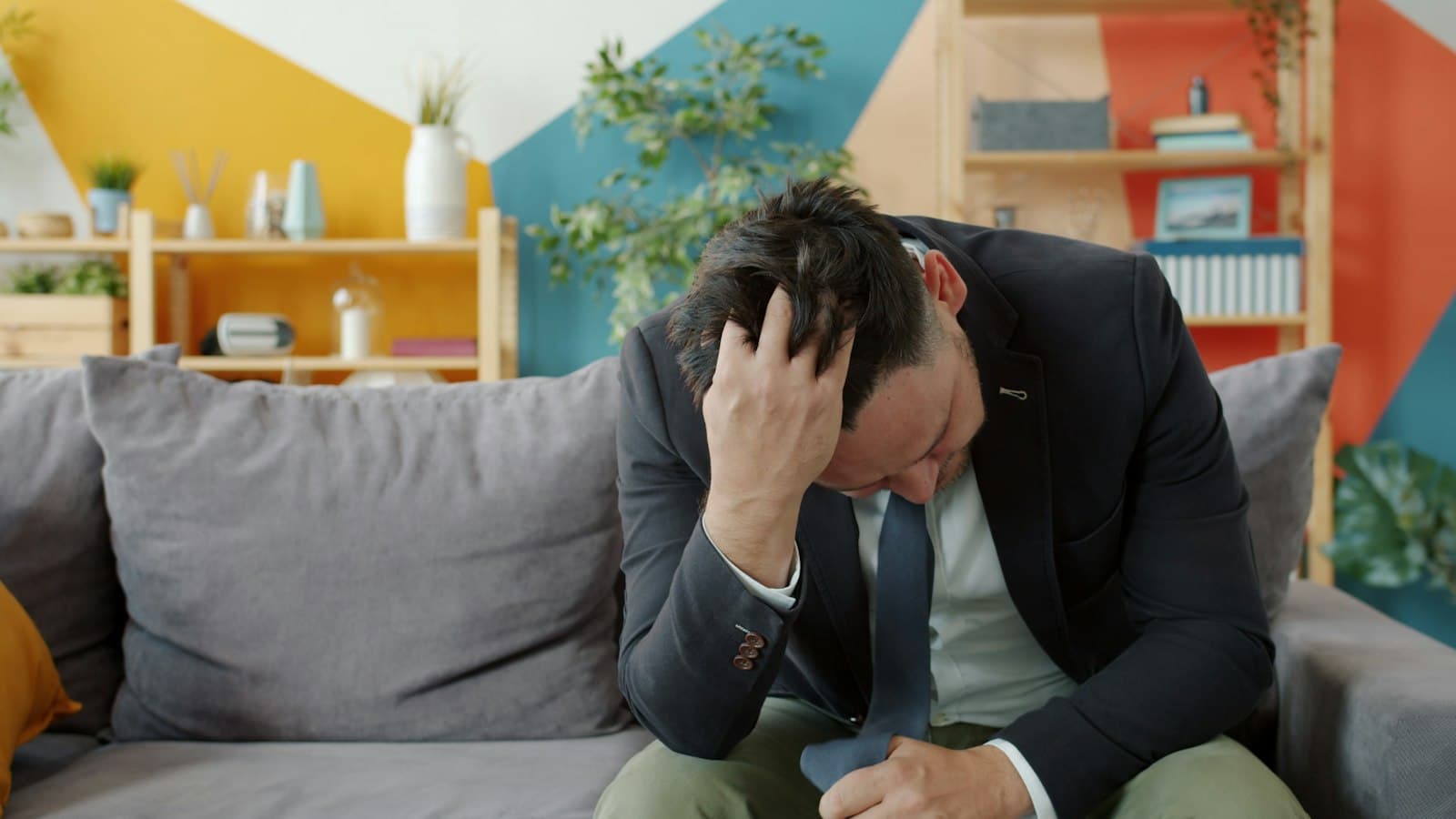 A man in a jacket sitting on a sofa with his head in his hand