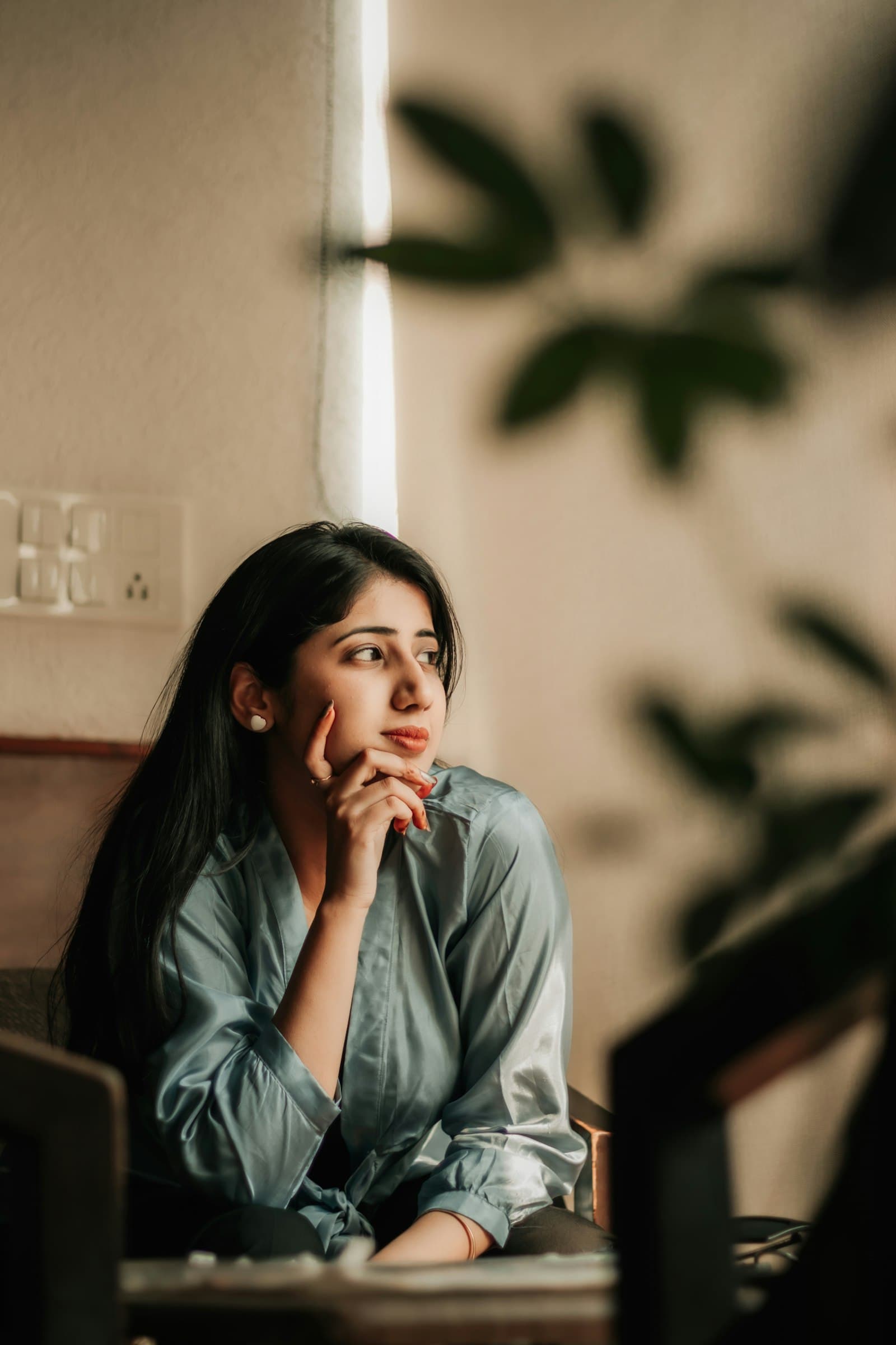A woman sitting by a window with her hand to her chin, looking off into the distance