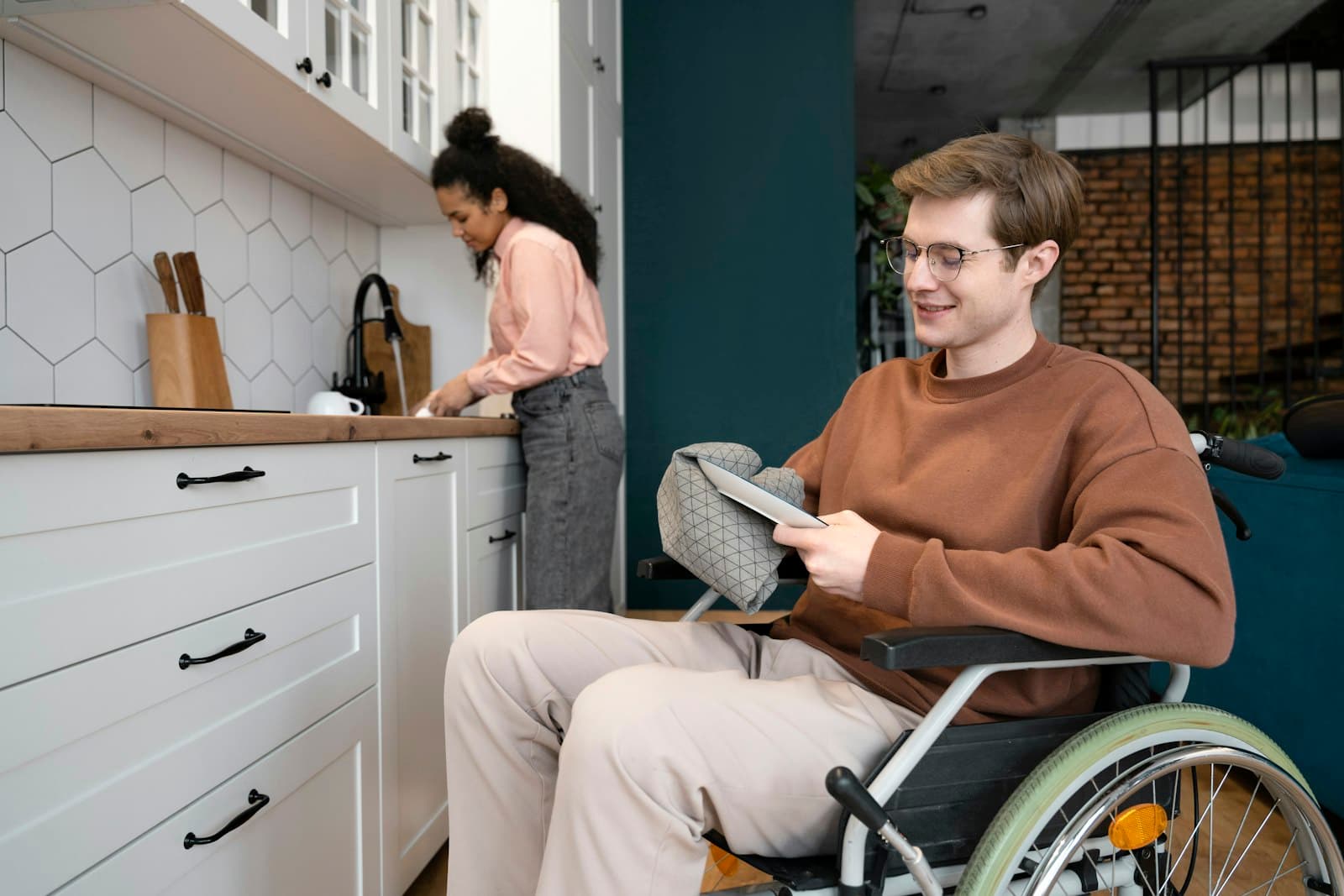 A young man in a wheelchair reading in a kitchen while his partner stands at the counter