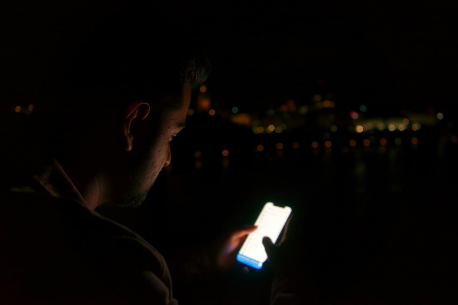 A man in the dark at night, his face lit by the glow of his phone screen