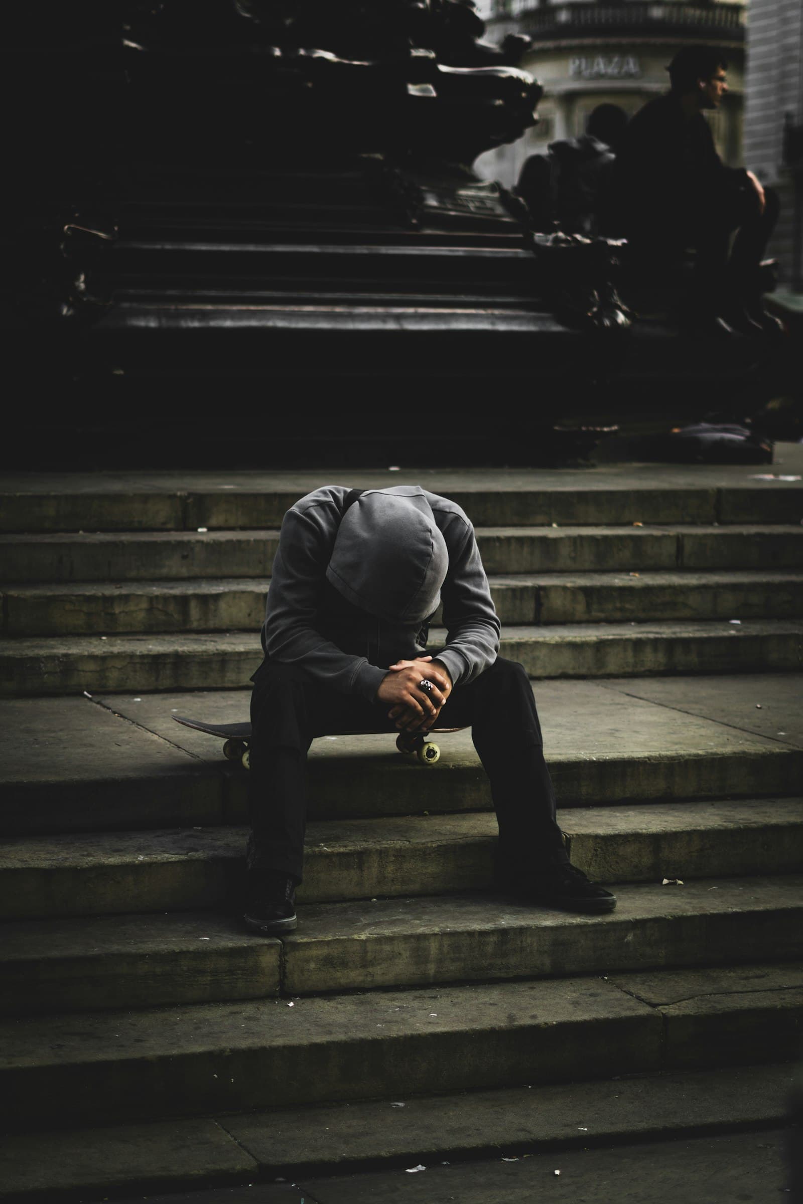 A man sitting alone on stone steps in dim light, head bowed under a cap