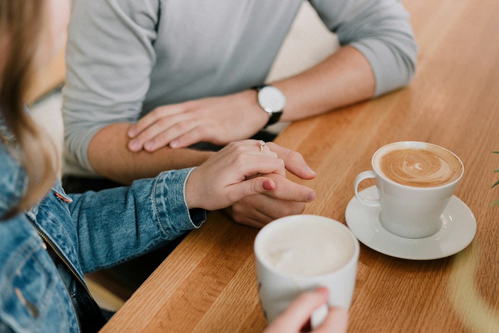 Two people at a wooden café table, one resting a hand gently on the other's beside cups of coffee