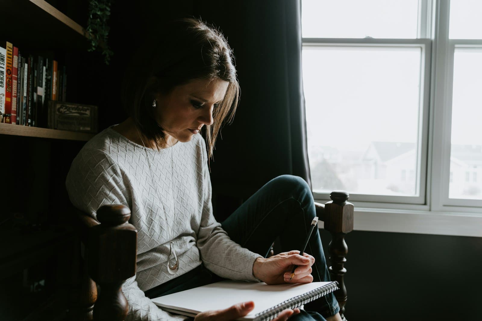 A woman sitting in a chair by a window, writing in a notebook