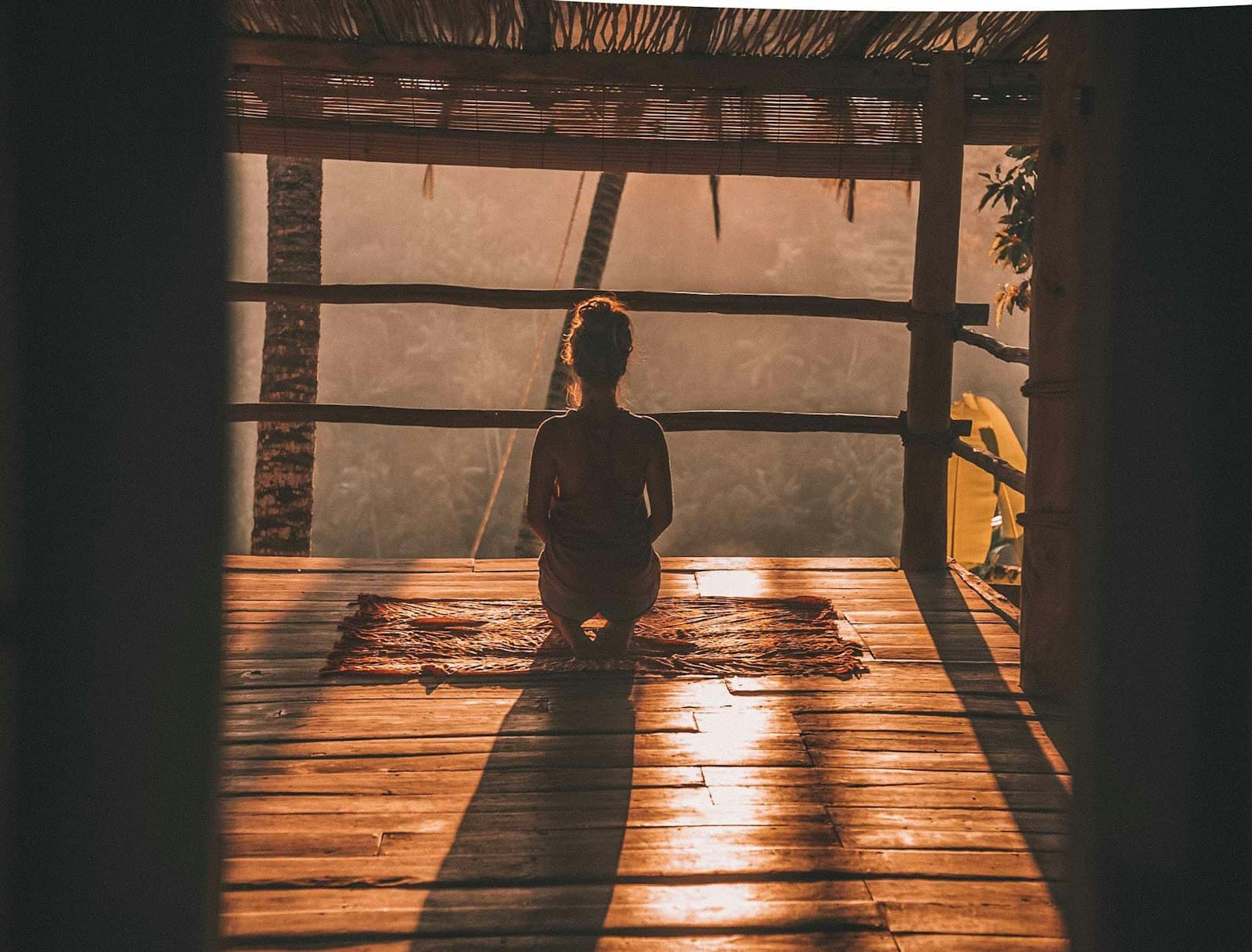 A woman sitting quietly on a wooden deck, silhouetted against warm morning light