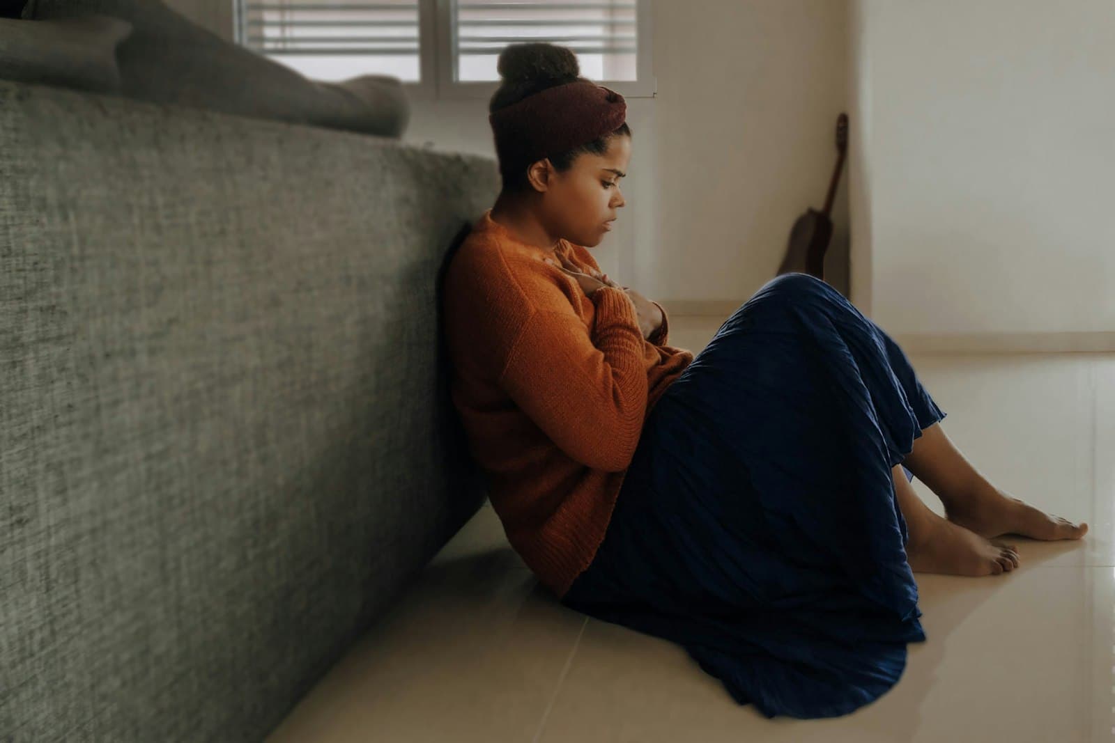 A woman in an orange sweater sitting on the floor against a sofa, arms folded, looking down