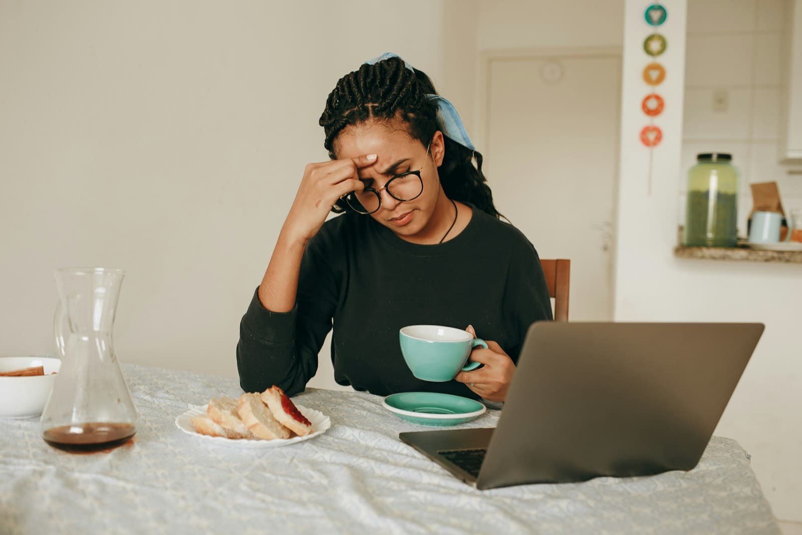 A woman in glasses at a kitchen table with a laptop and tea, rubbing her forehead