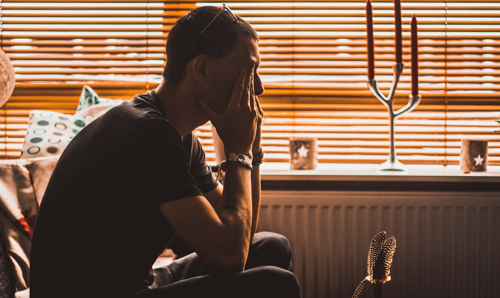A man sitting by a window with blinds, rubbing his eyes with his hand