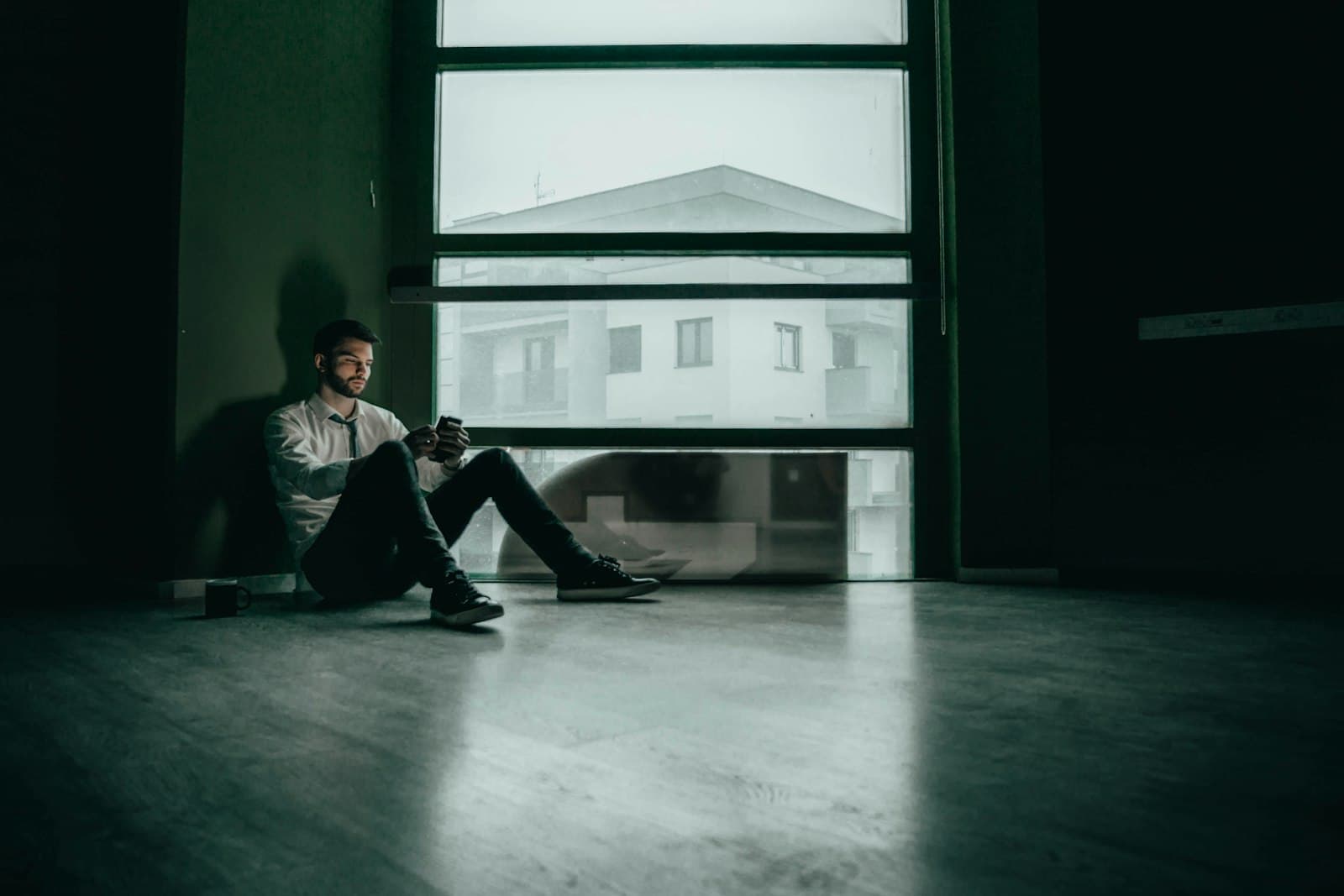 A man sitting alone on the floor of an empty room by a large window, looking at his phone