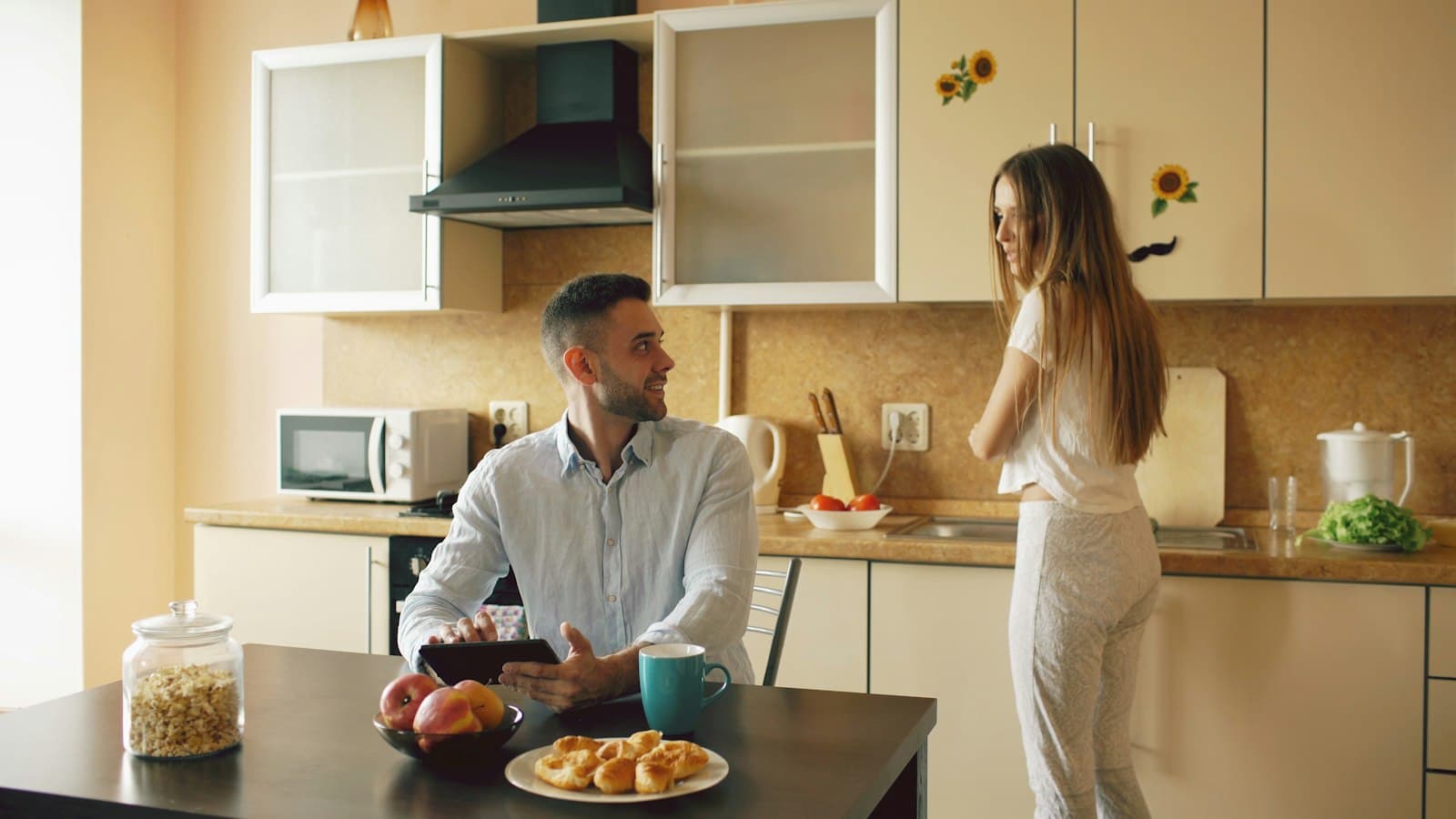 A man sitting at a kitchen table while a woman stands at the counter across the room