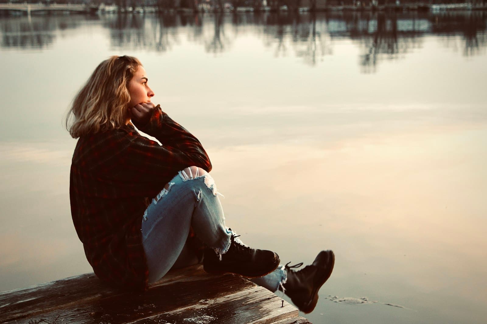 A woman in a dark jacket sitting by still water, resting her chin on her hand