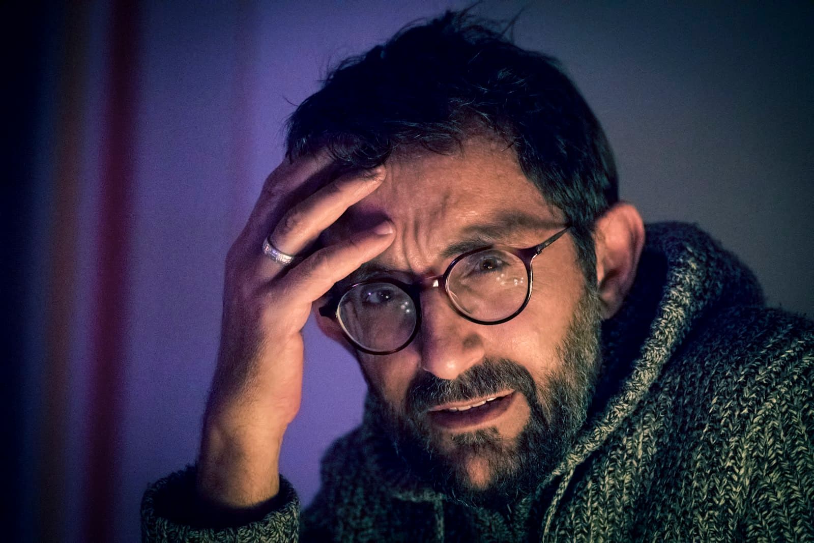 A bearded man in round glasses holding a hand to his head, looking worried