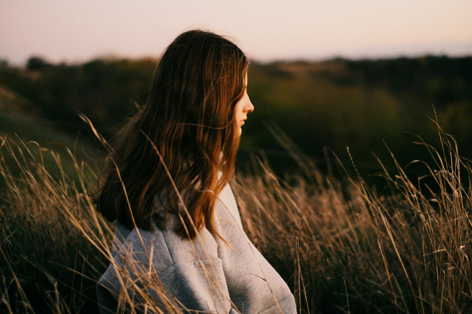 A woman standing in tall grass at dusk, looking out across the hills
