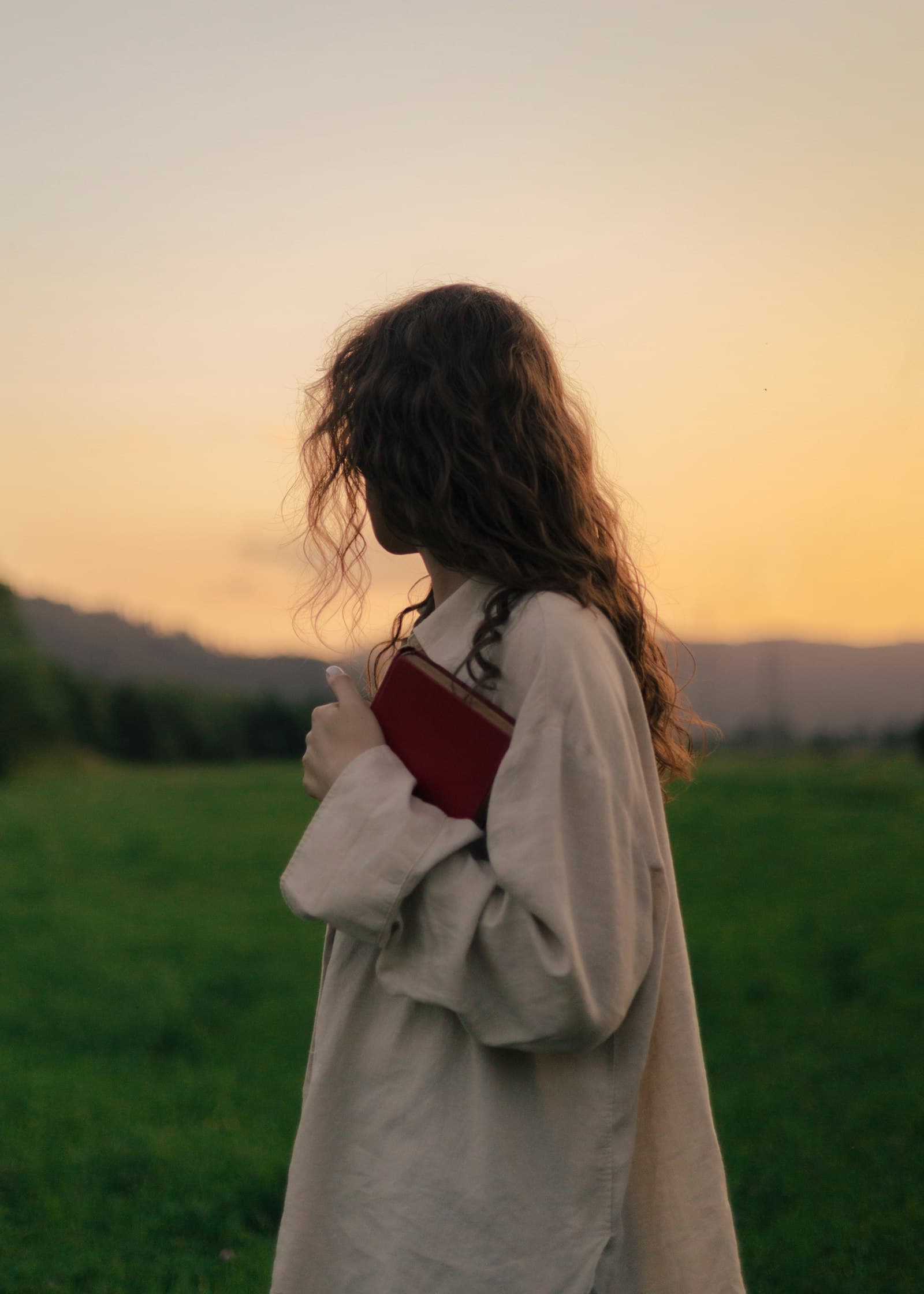 A woman in a long cardigan standing in a field at dusk, holding a book to her chest