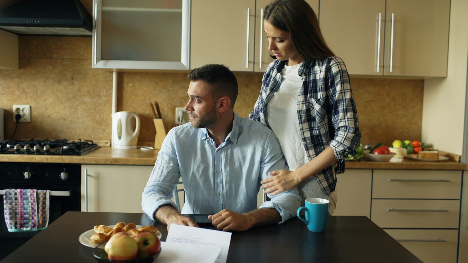 A woman with her hand on her partner's shoulder as he sits at the kitchen table with paperwork