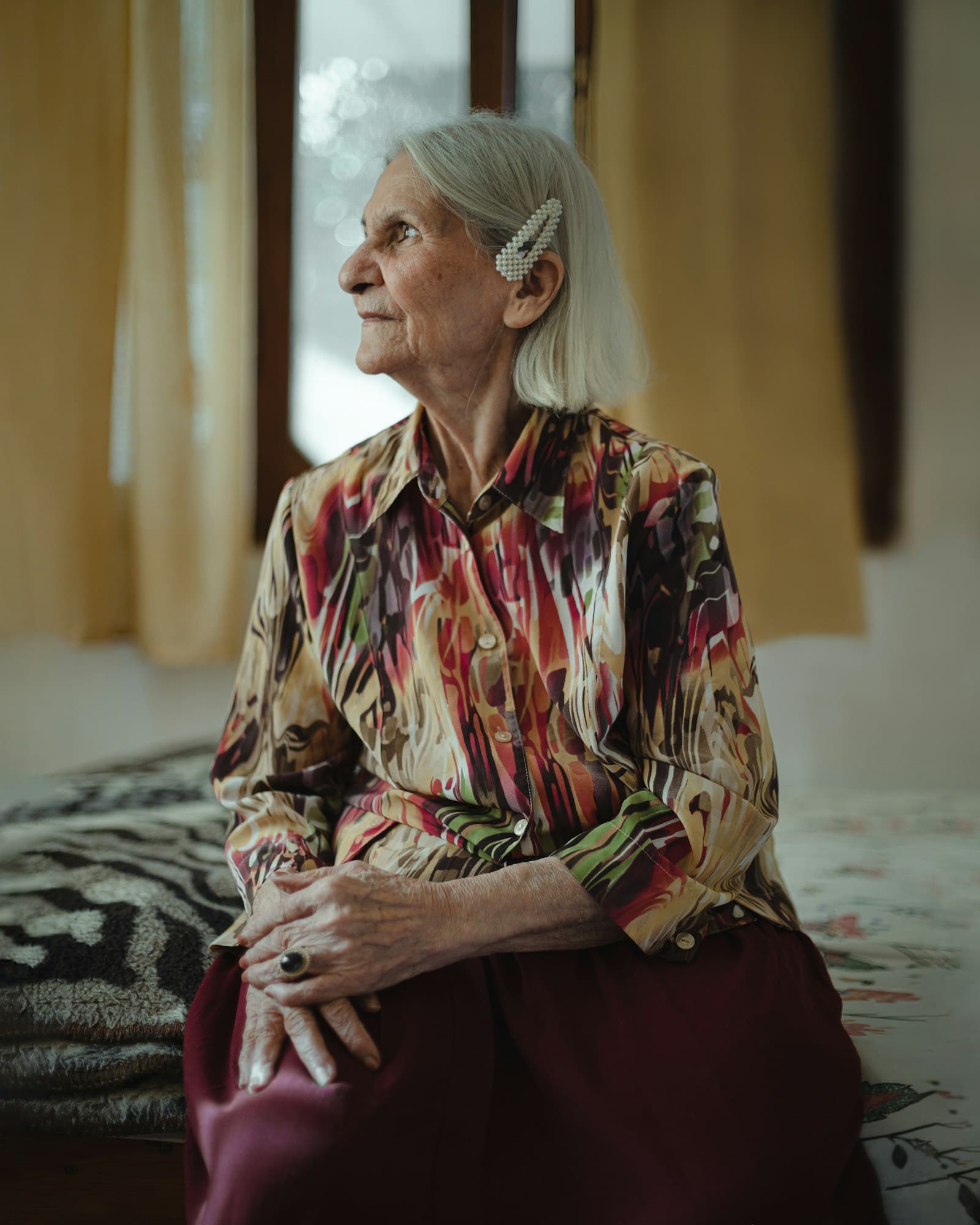 An elderly woman with white hair sitting on the edge of a bed, looking toward the window