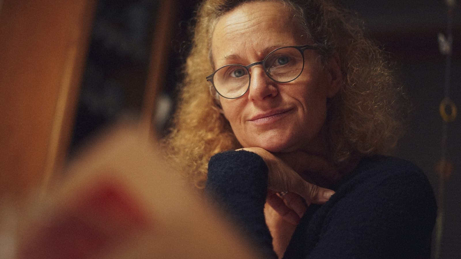 A middle-aged woman with curly hair and glasses resting her chin on her hand