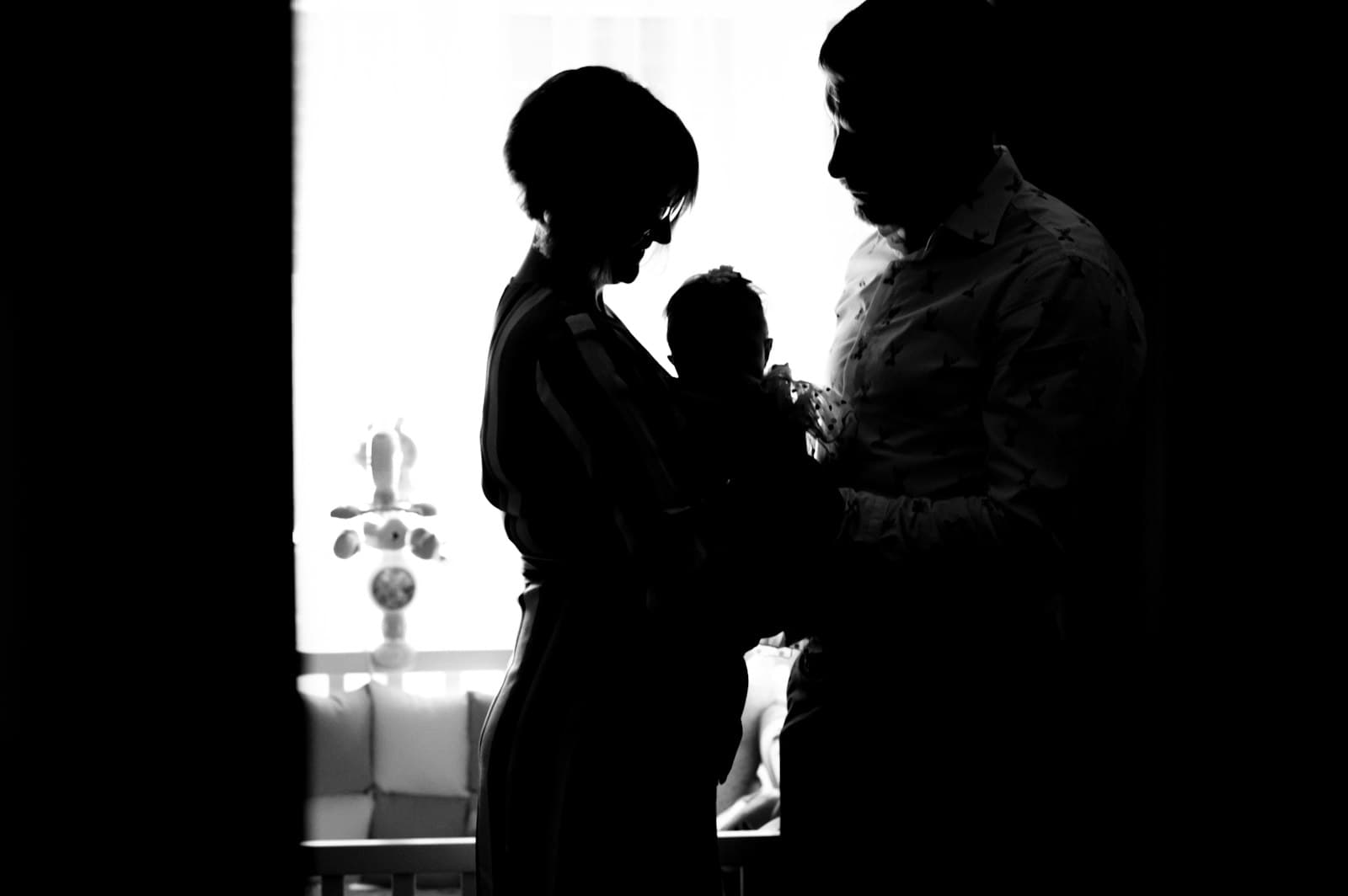 Silhouettes of two parents holding a newborn between them by a bright window