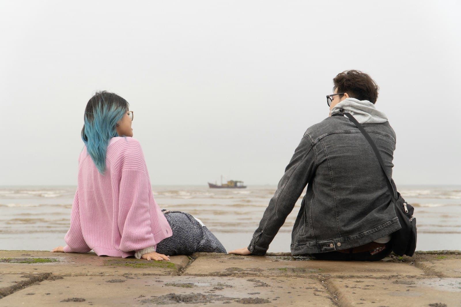 A man and a woman sitting apart on a stone ledge by the sea, both looking toward the water