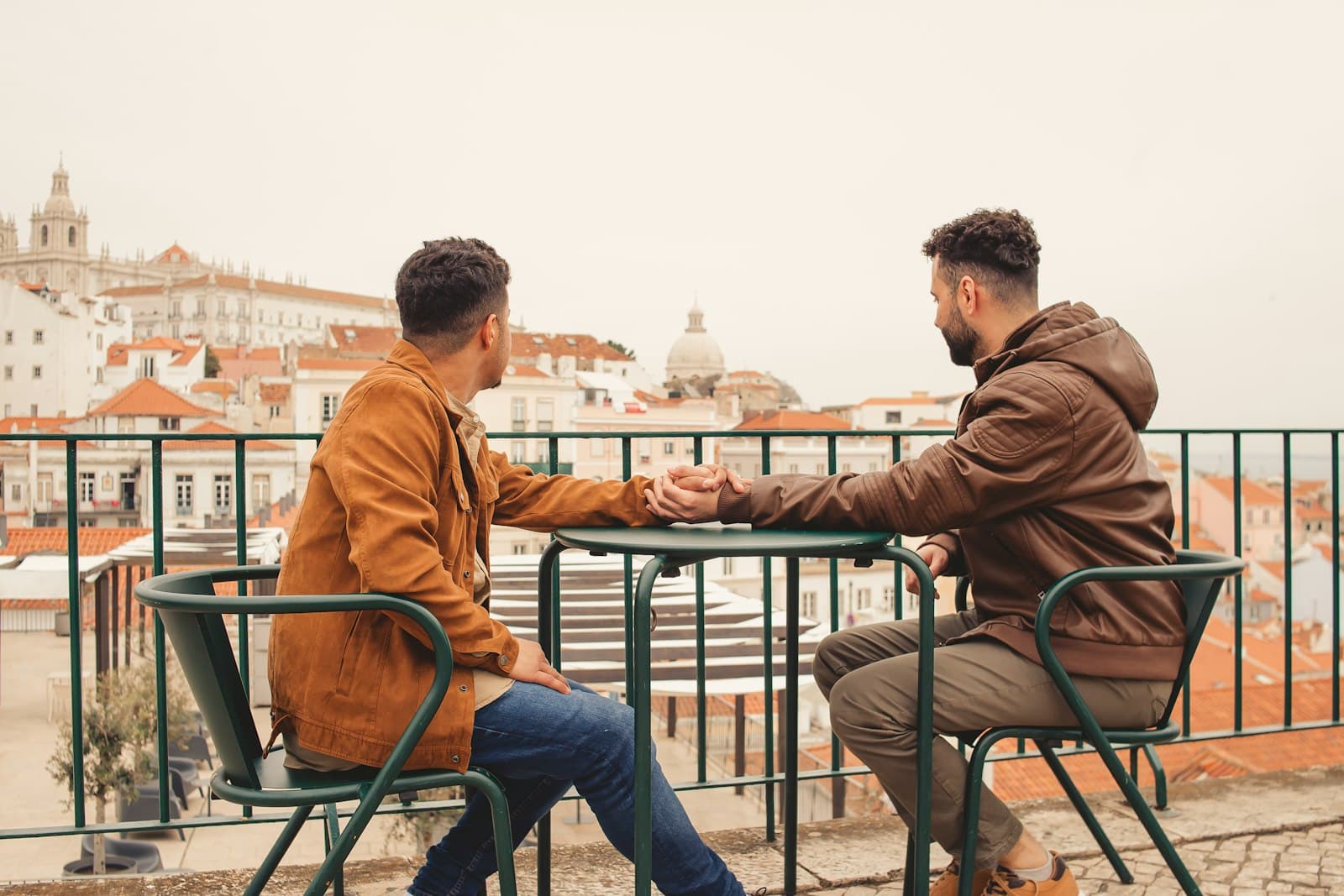 Two men holding hands across a small table on a rooftop terrace overlooking the city