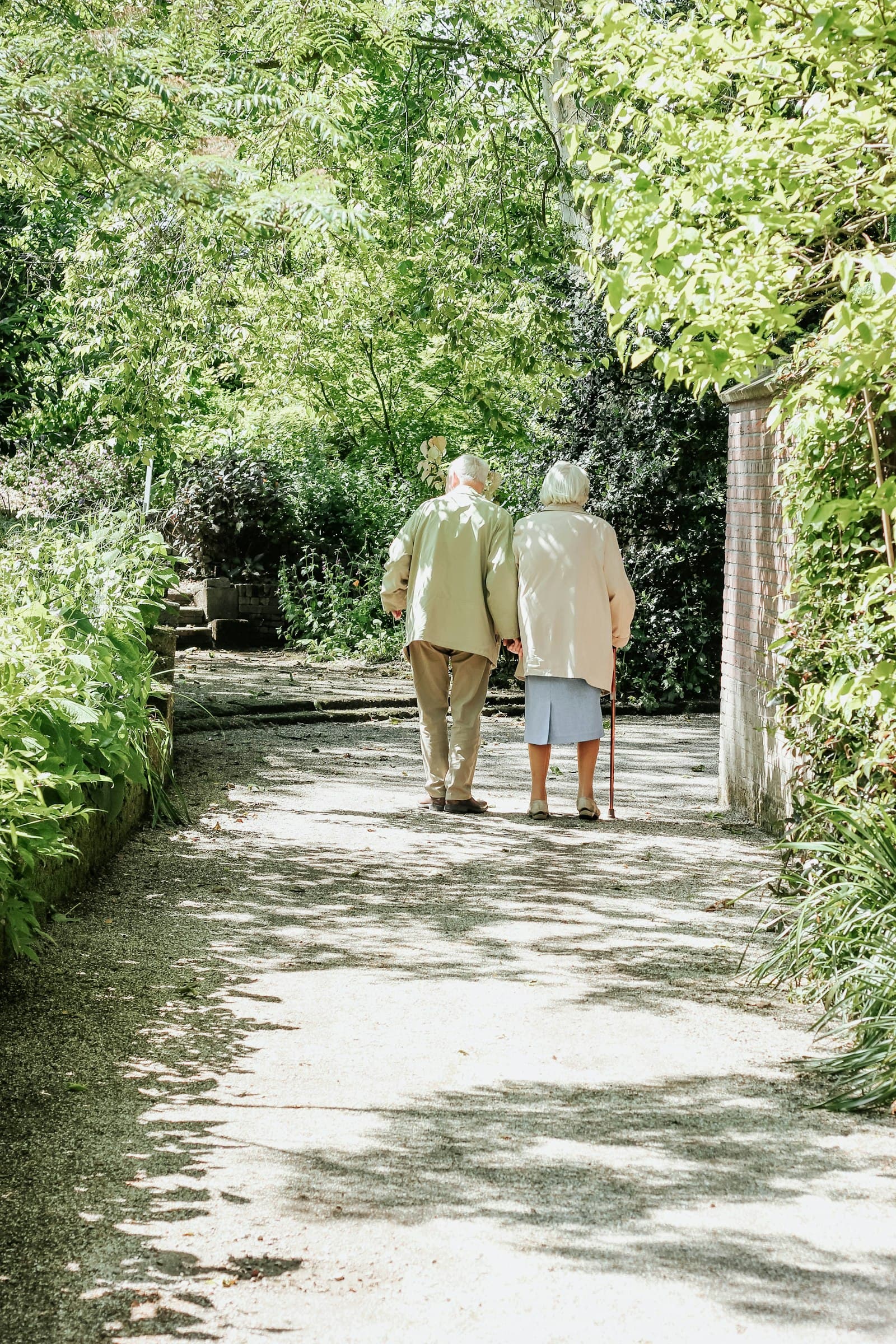 An elderly couple walking side by side down a leafy garden path