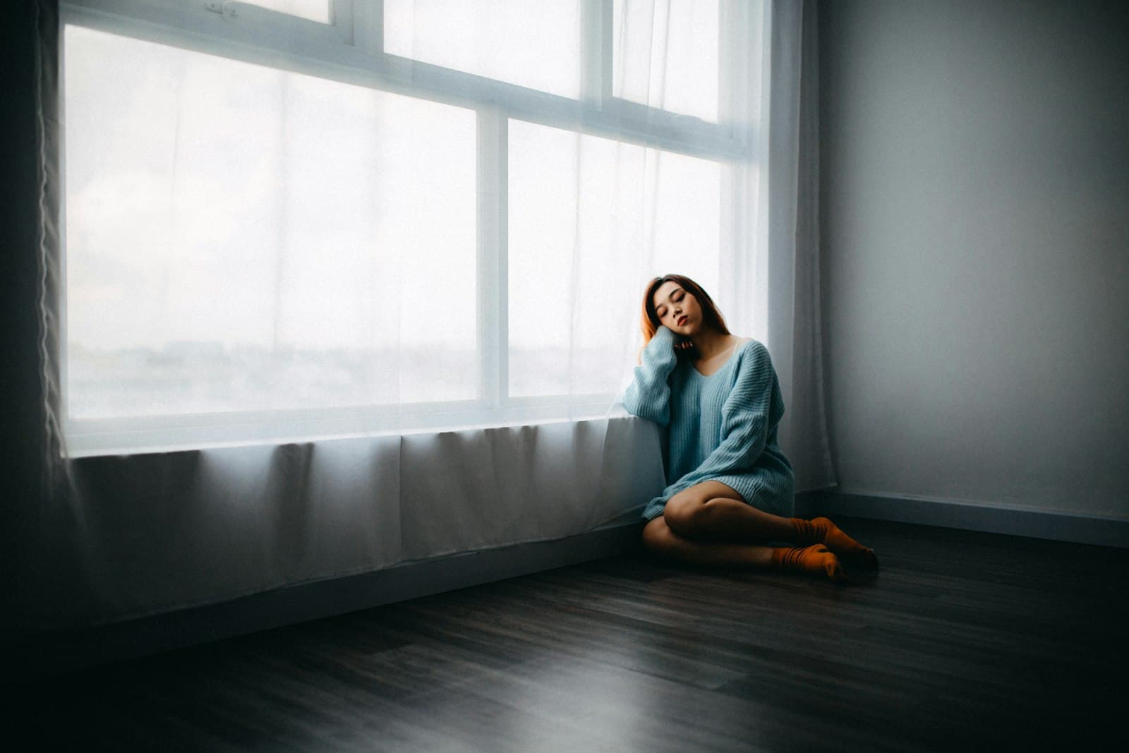 A woman in a sweater sitting alone on the floor beside a bright, curtained window
