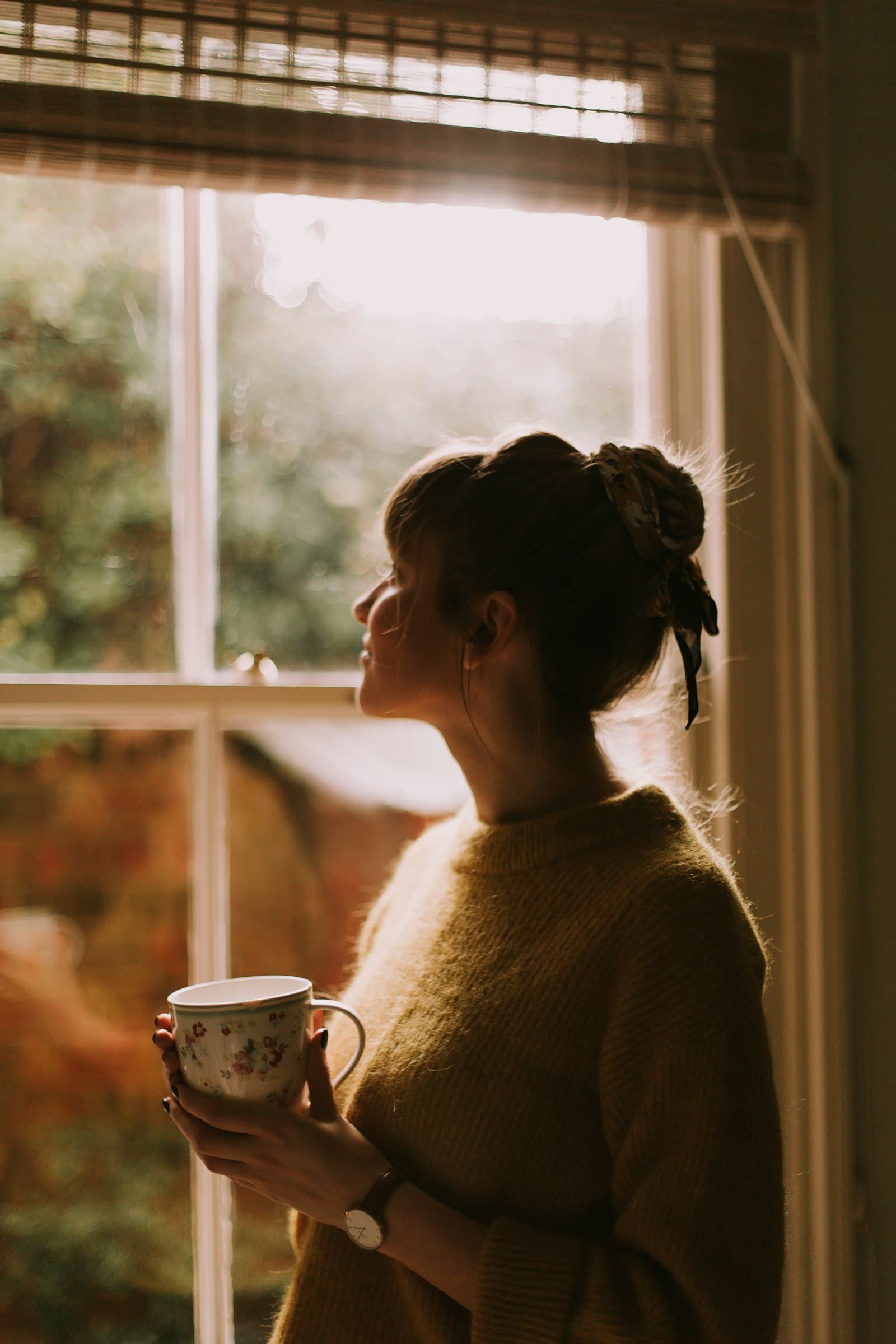 A woman holding a mug by a sunlit window, looking out thoughtfully