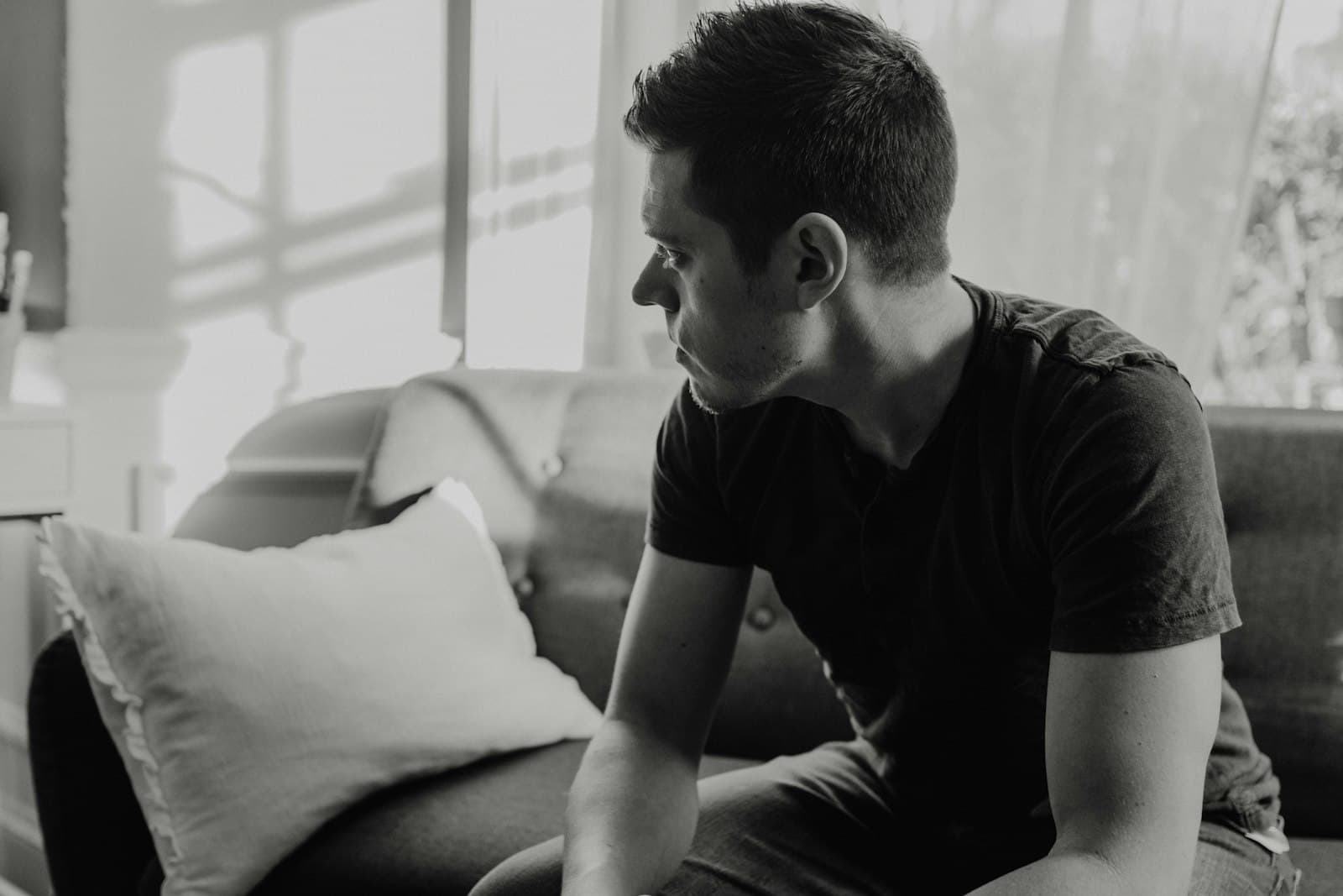 A man in a dark T-shirt sitting on a sofa, looking off to the side in thought, in black and white