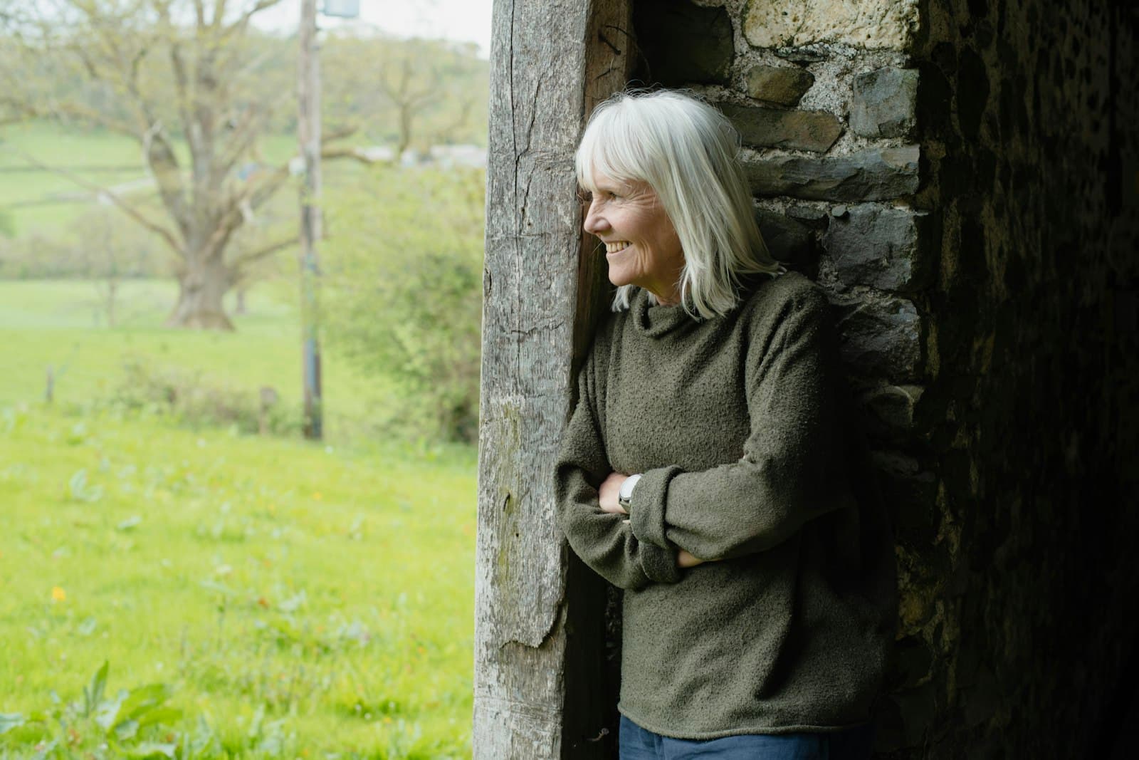 A smiling woman with grey hair and a green sweater leaning in a stone doorway beside a field
