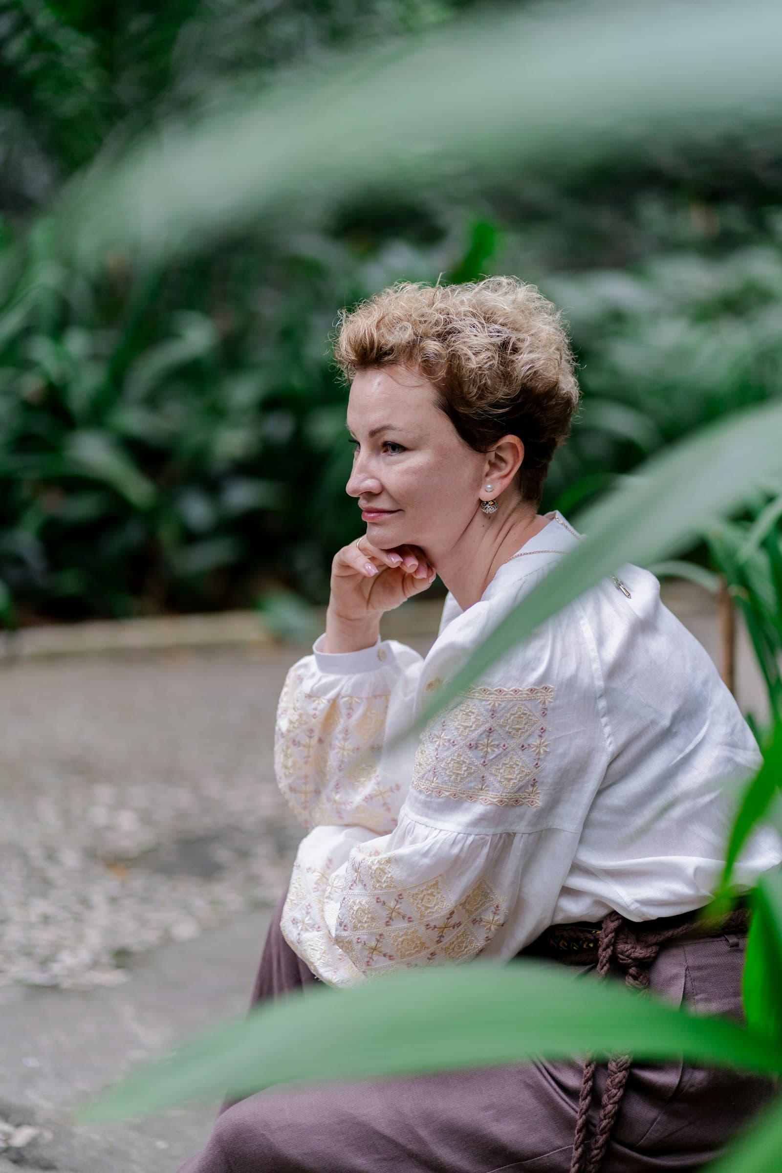 A woman in her fifties in a white blouse sitting in a garden, resting her chin on her hand
