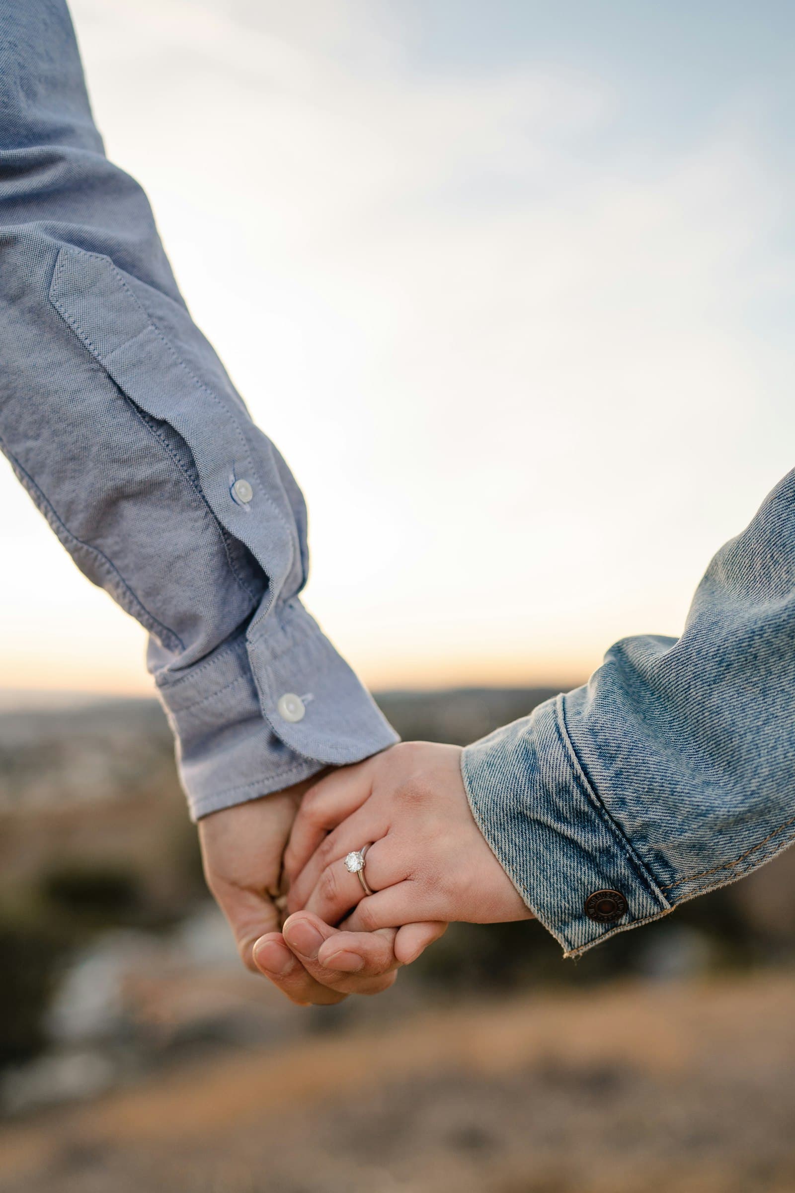 Close-up of two people holding hands outdoors, one wearing a ring and a denim jacket