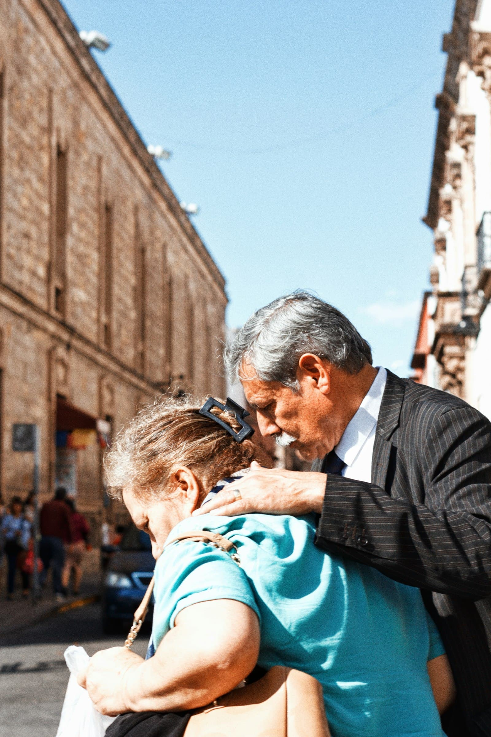 An older man in a suit hugging a woman and kissing the top of her head on a sunny street