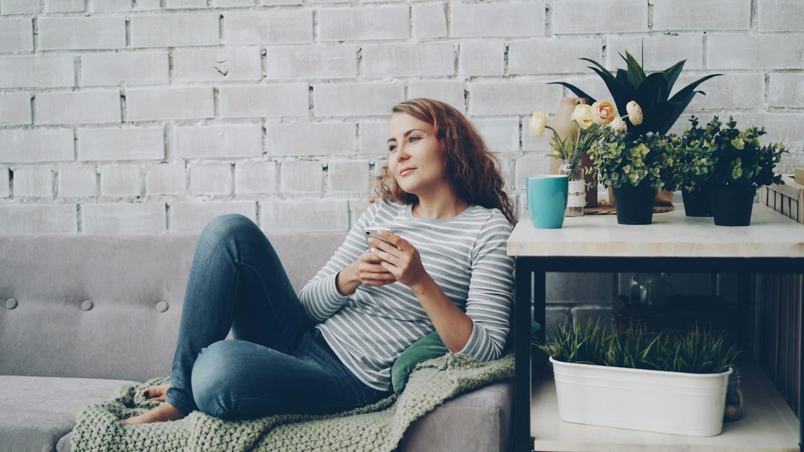 A woman resting on a grey sofa with a mug, gazing out of the frame