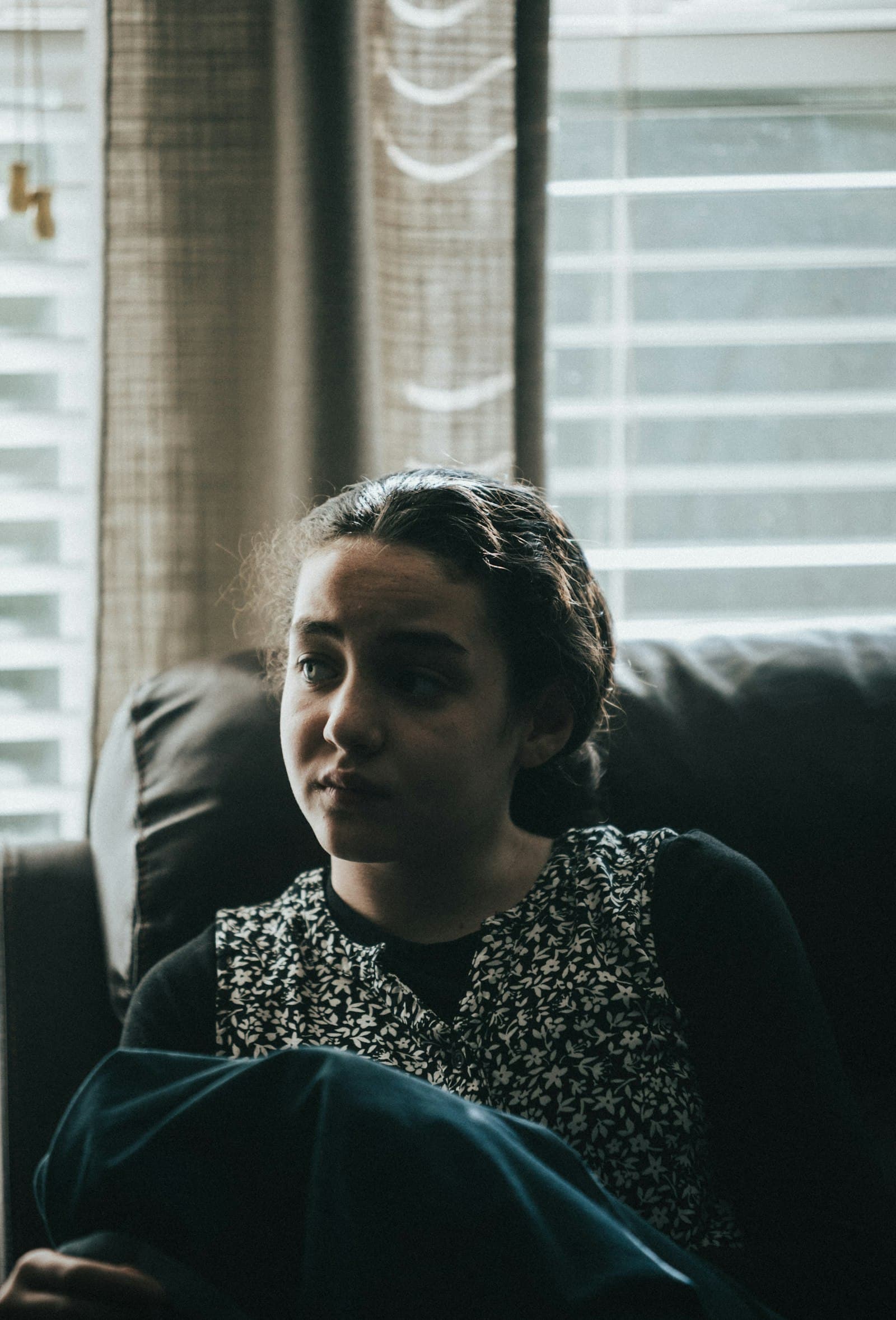A woman sitting in a dark armchair by a window, gazing off to the side