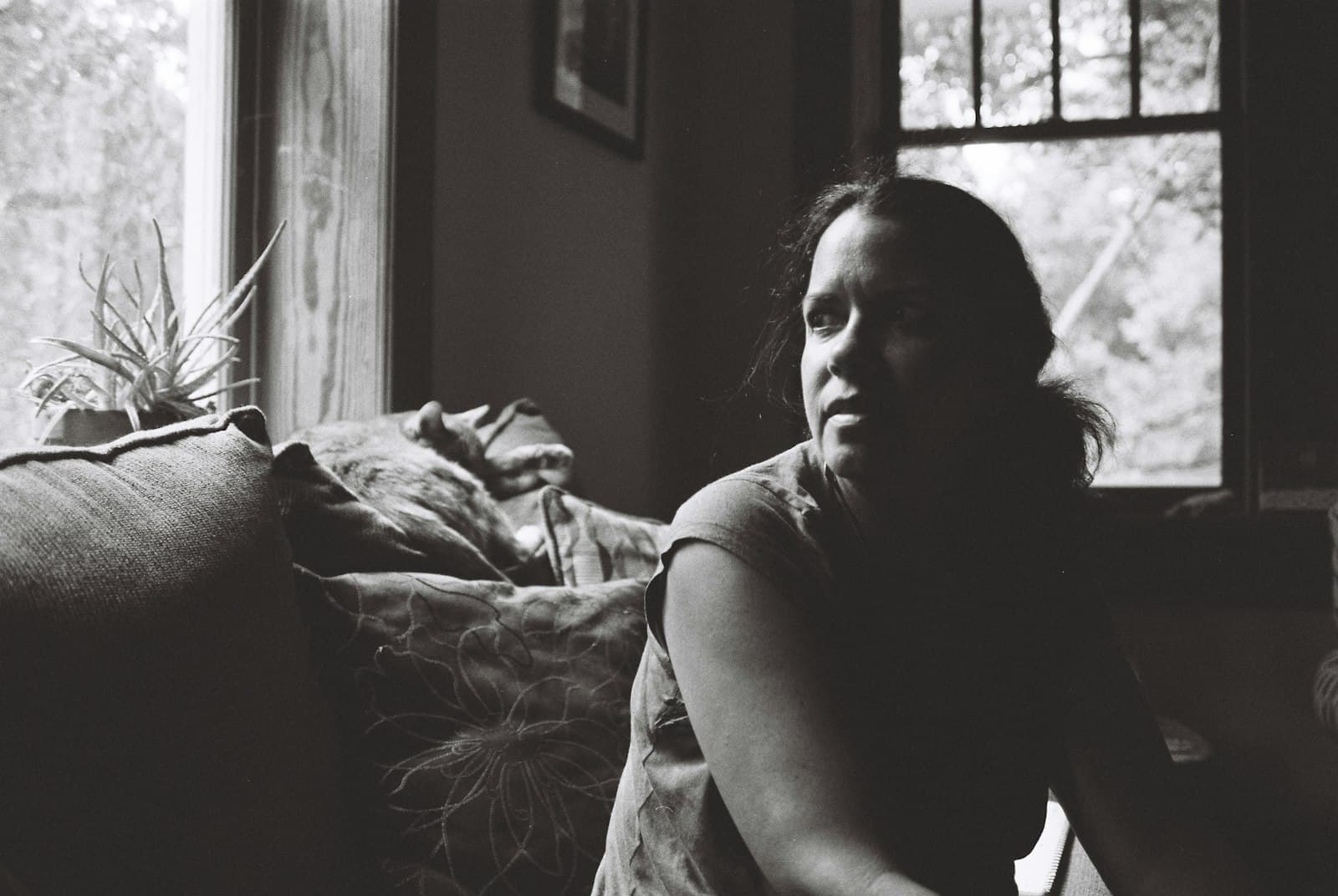 A woman sitting alone on a sofa in a dim room, looking toward the window, in black and white