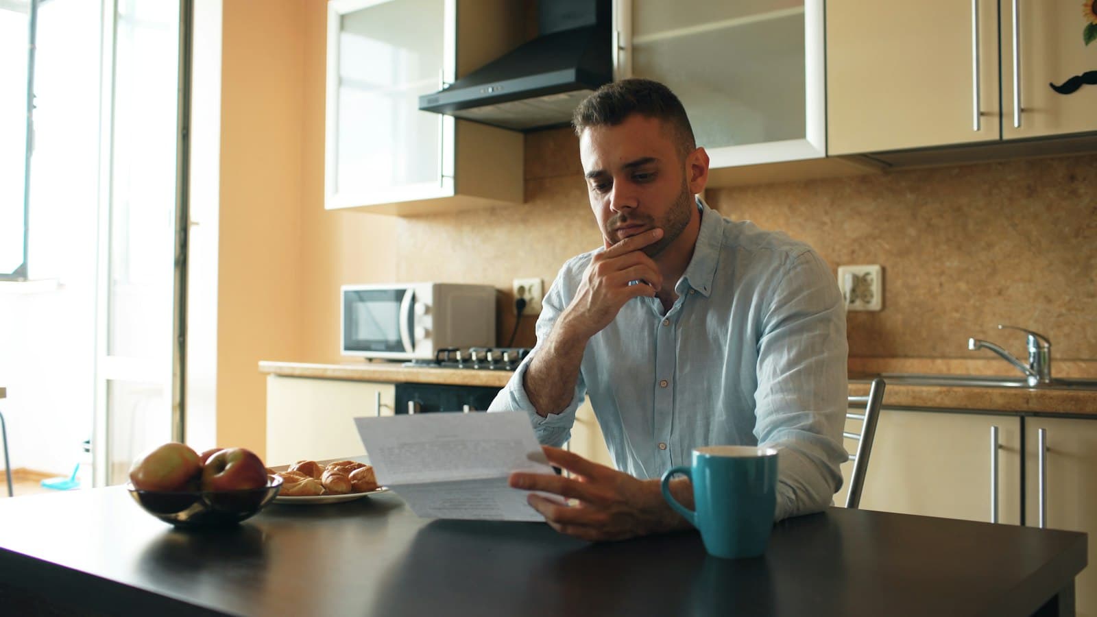 A man in a light blue shirt reading a letter at his kitchen table, hand at his chin