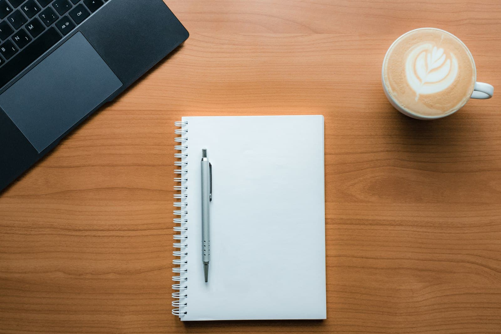 A blank spiral notepad and a pen on a wooden desk beside a laptop and a latte