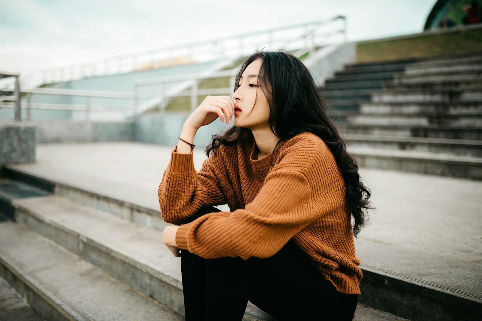 A woman in an orange sweater sitting on outdoor steps, chin in hand, deep in thought