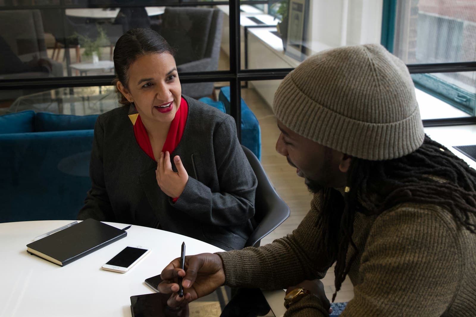 A woman and a man in a knit cap talking at a table, a notebook between them