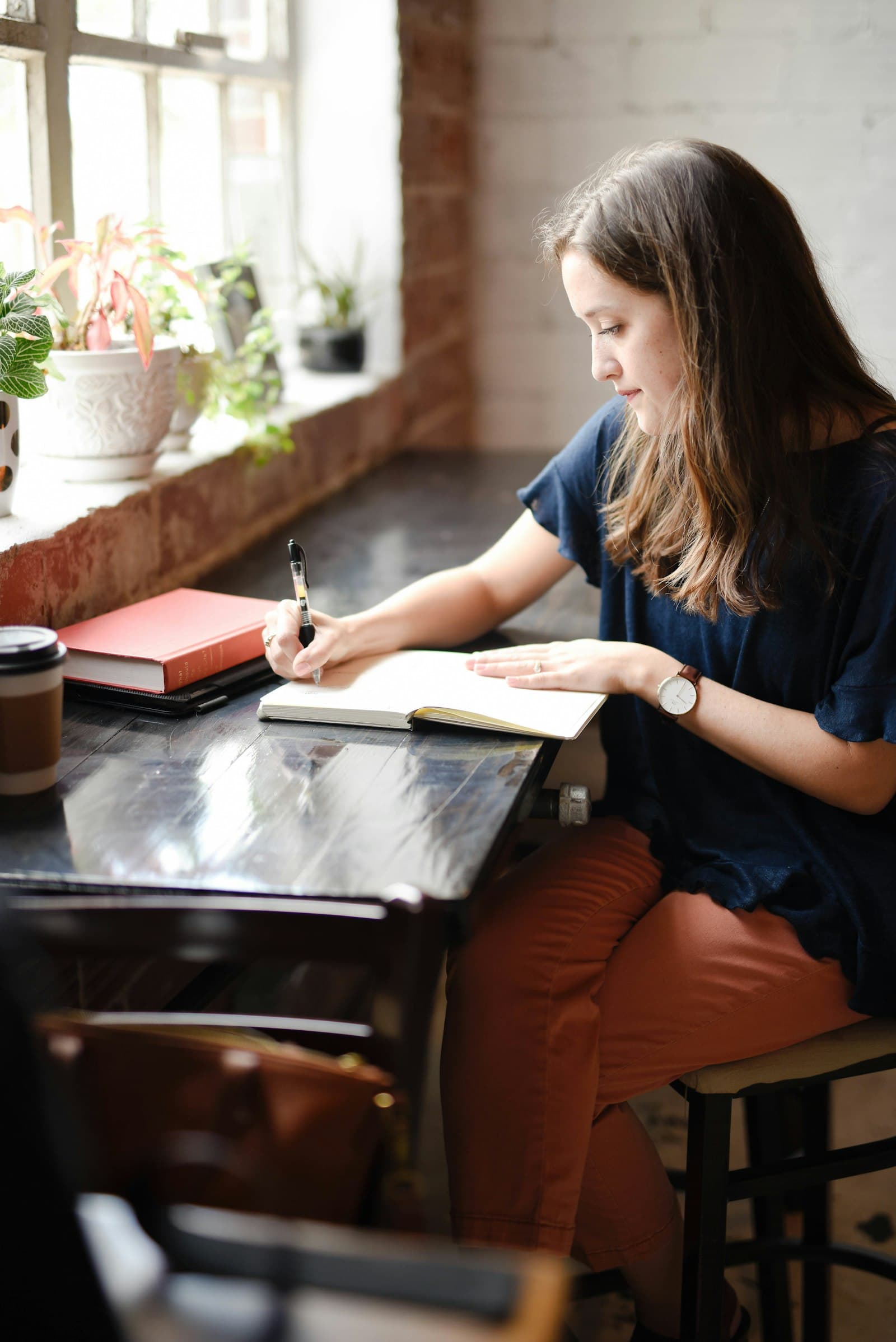 A woman writing in a notebook at a small table by a bright café window