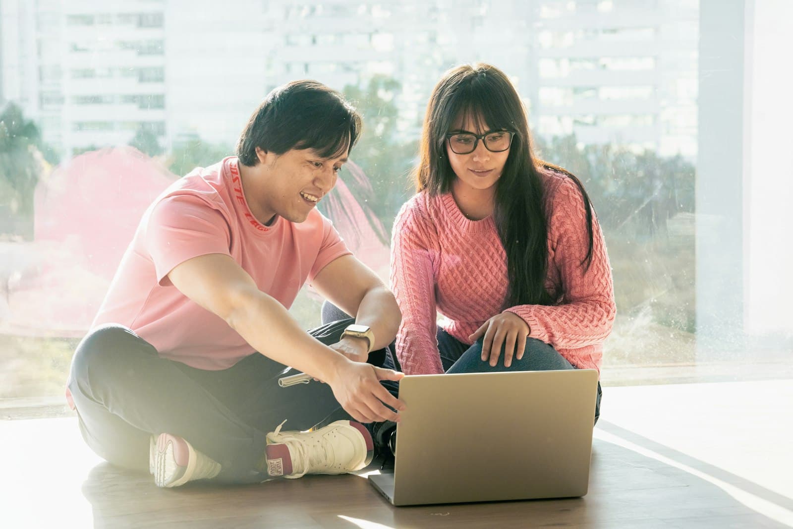 A couple in pink sweaters sitting on the floor by a bright window, looking at a laptop together